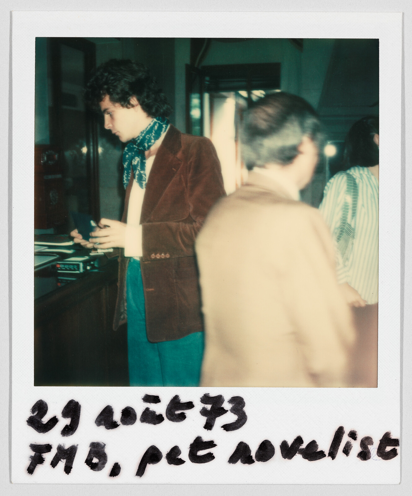 A young man in a brown jacket stands at a counter. Handwritten text reads: "29 août 73, FMB, pet novelist."