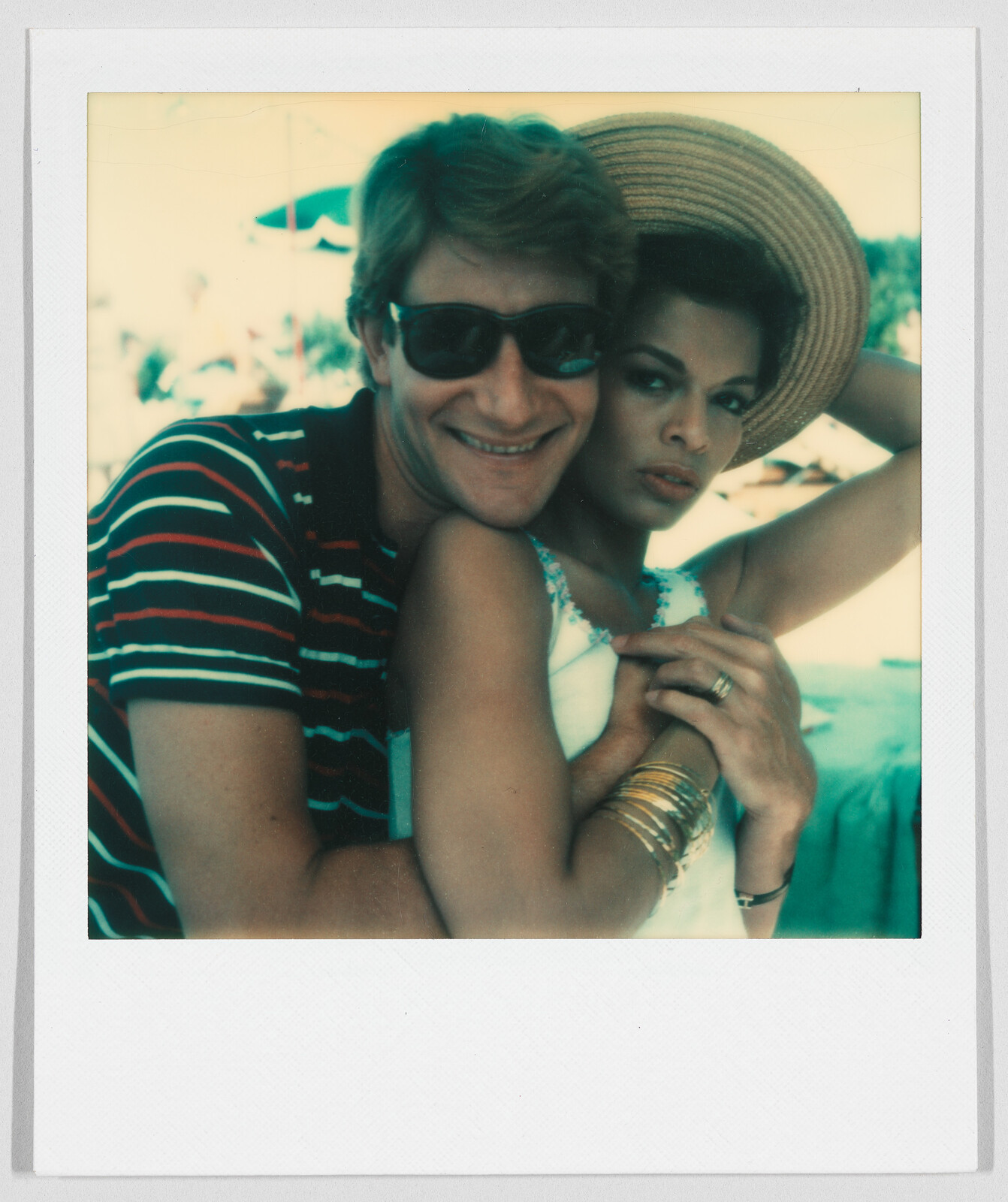 A man in sunglasses hugs a woman wearing a sunhat and bracelets; both are looking at the camera.