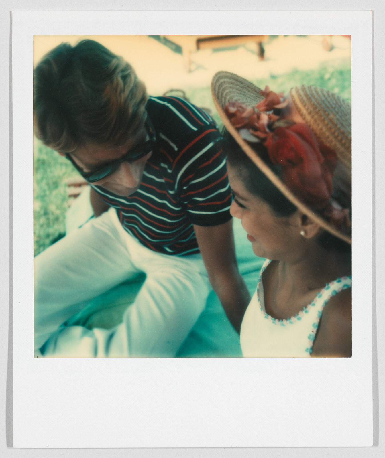 Two people sitting close together outdoors, smiling and talking; one wears sunglasses, the other a straw hat.