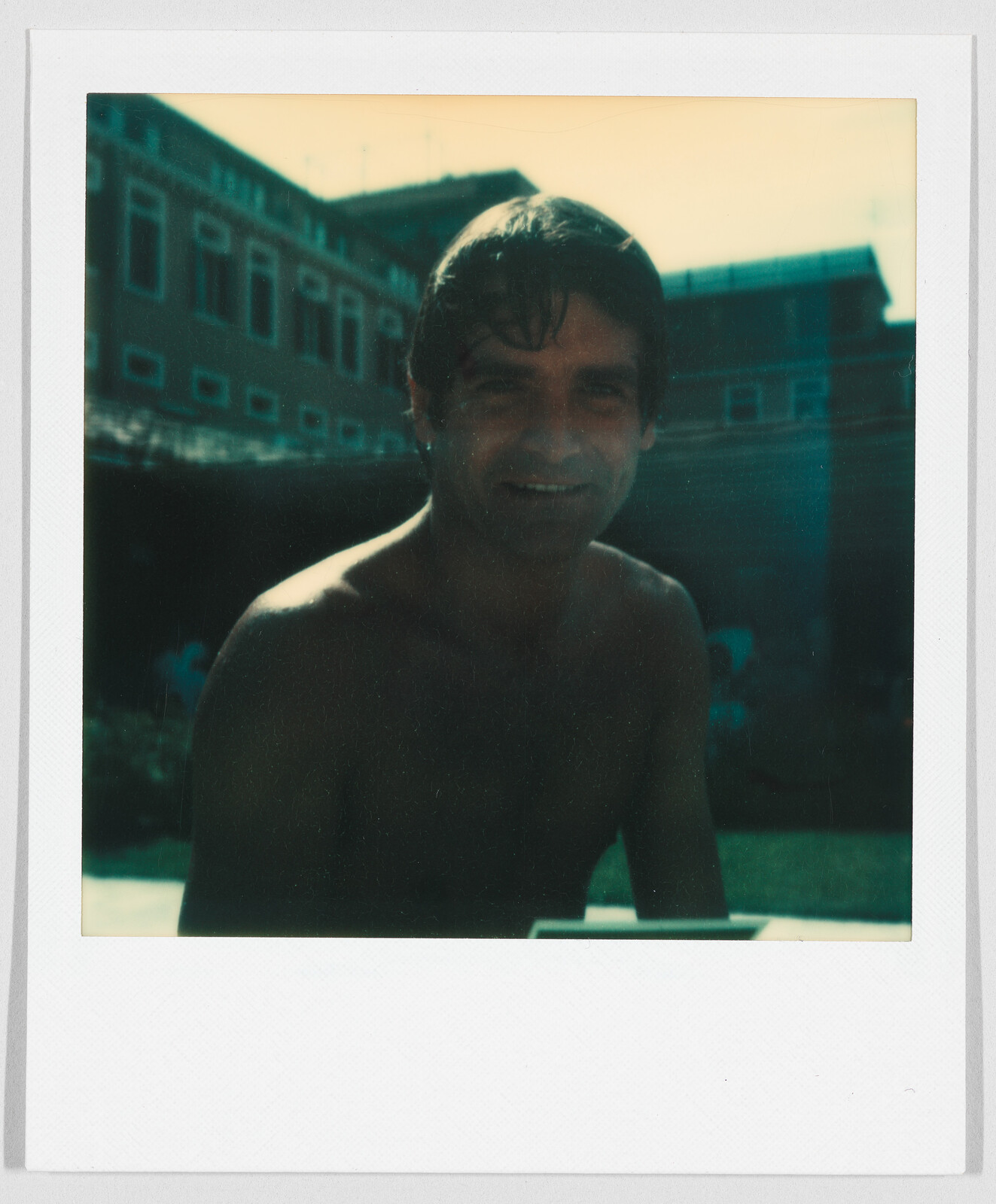 A man with wet hair and no shirt smiles outdoors, with buildings visible in the background.