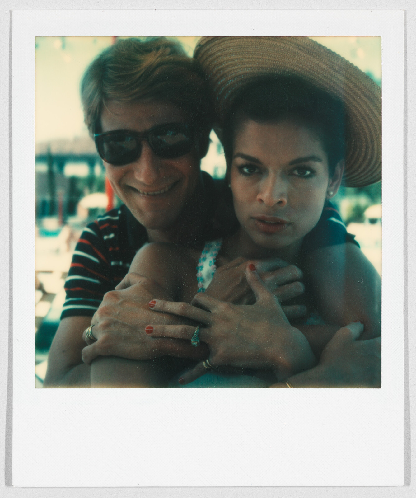 A man wearing sunglasses hugs a woman with a sunhat from behind; both look toward the camera.