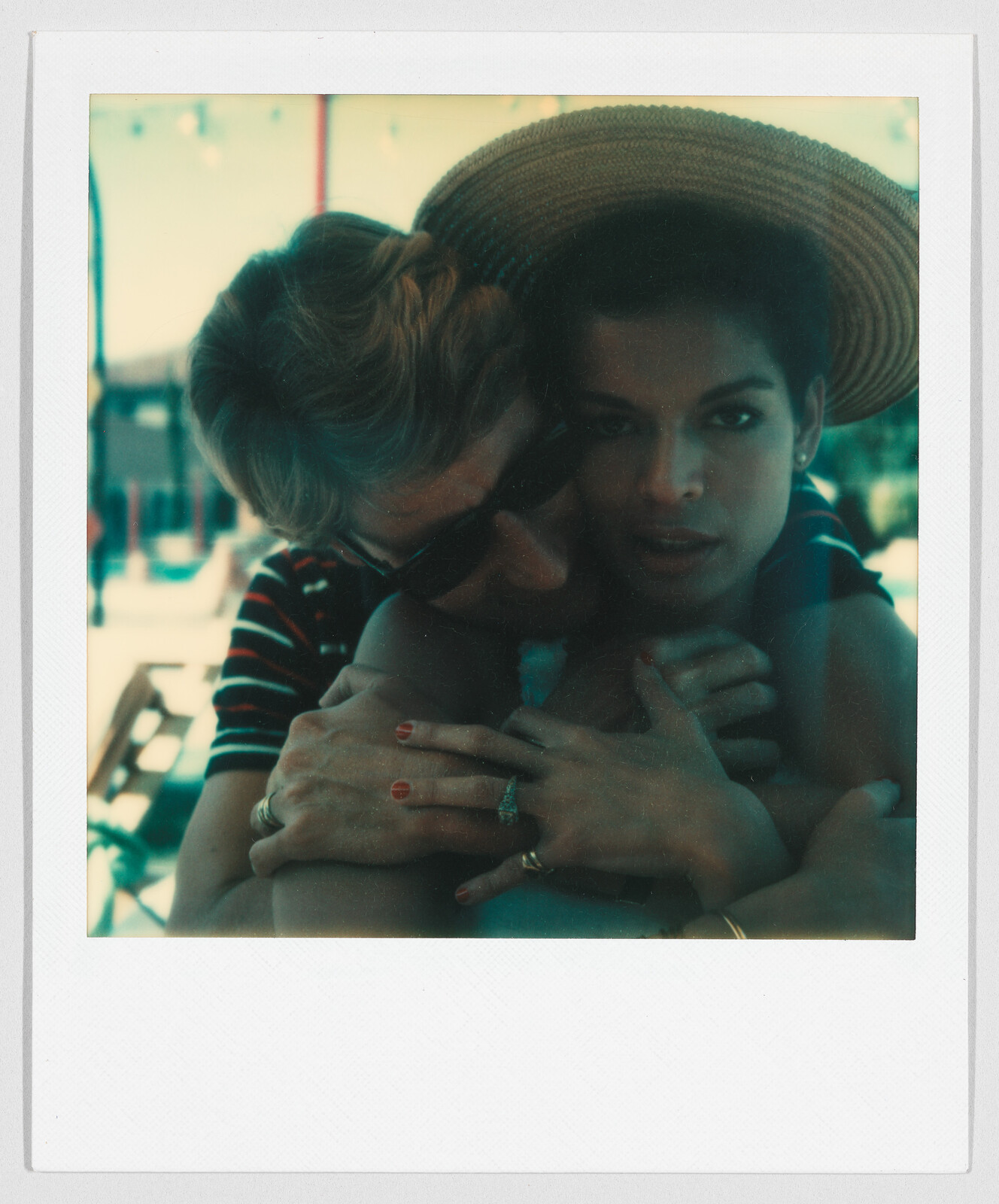 Two people embrace closely outdoors; one wears sunglasses and the other has a wide-brimmed hat.