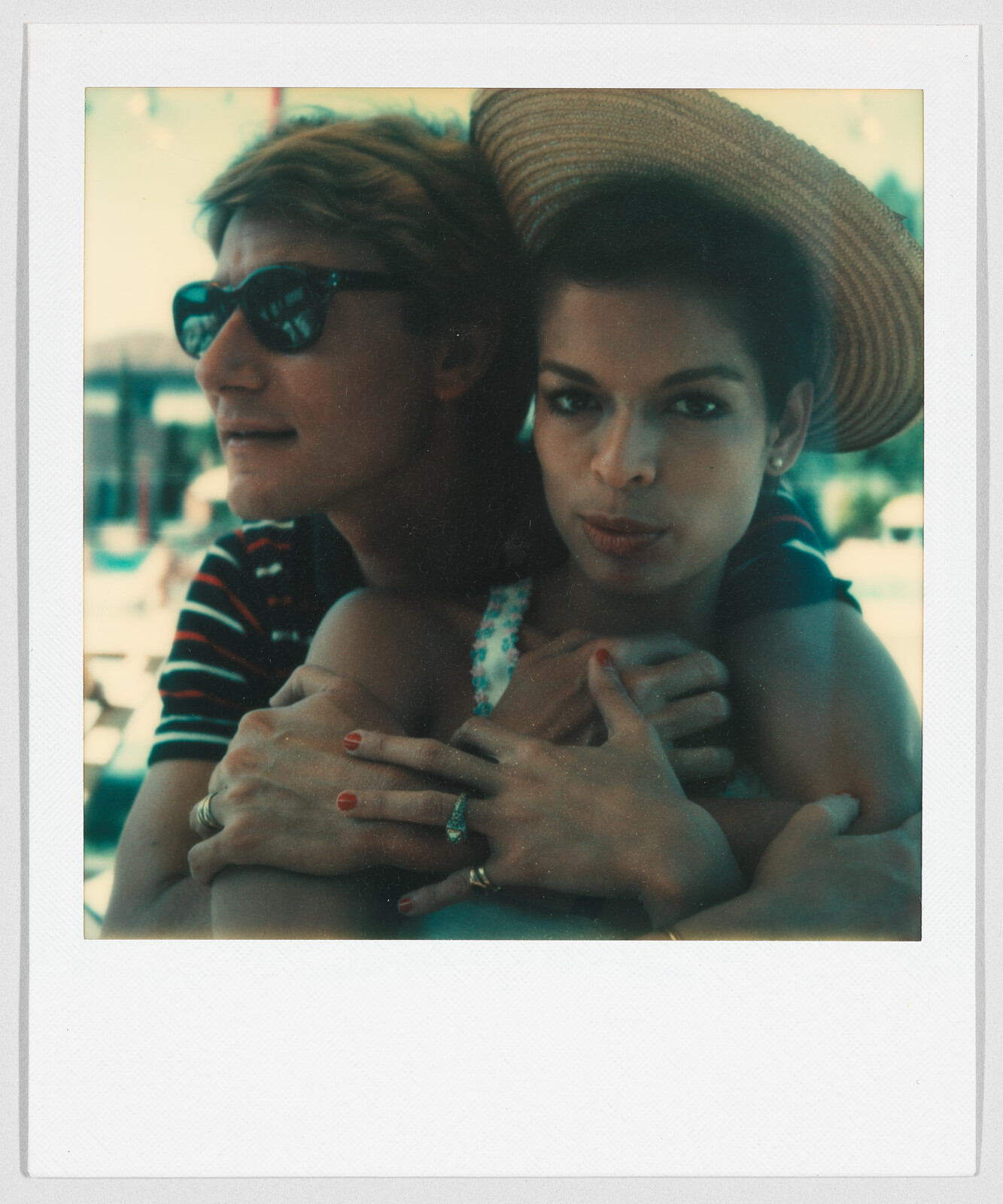 Two people sit close together outdoors; one wears sunglasses and the other has a wide-brimmed hat and red nails.