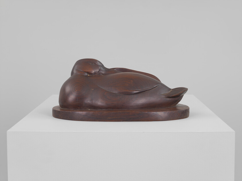A smooth wooden sculpture of a sleeping duck rests on a white pedestal.