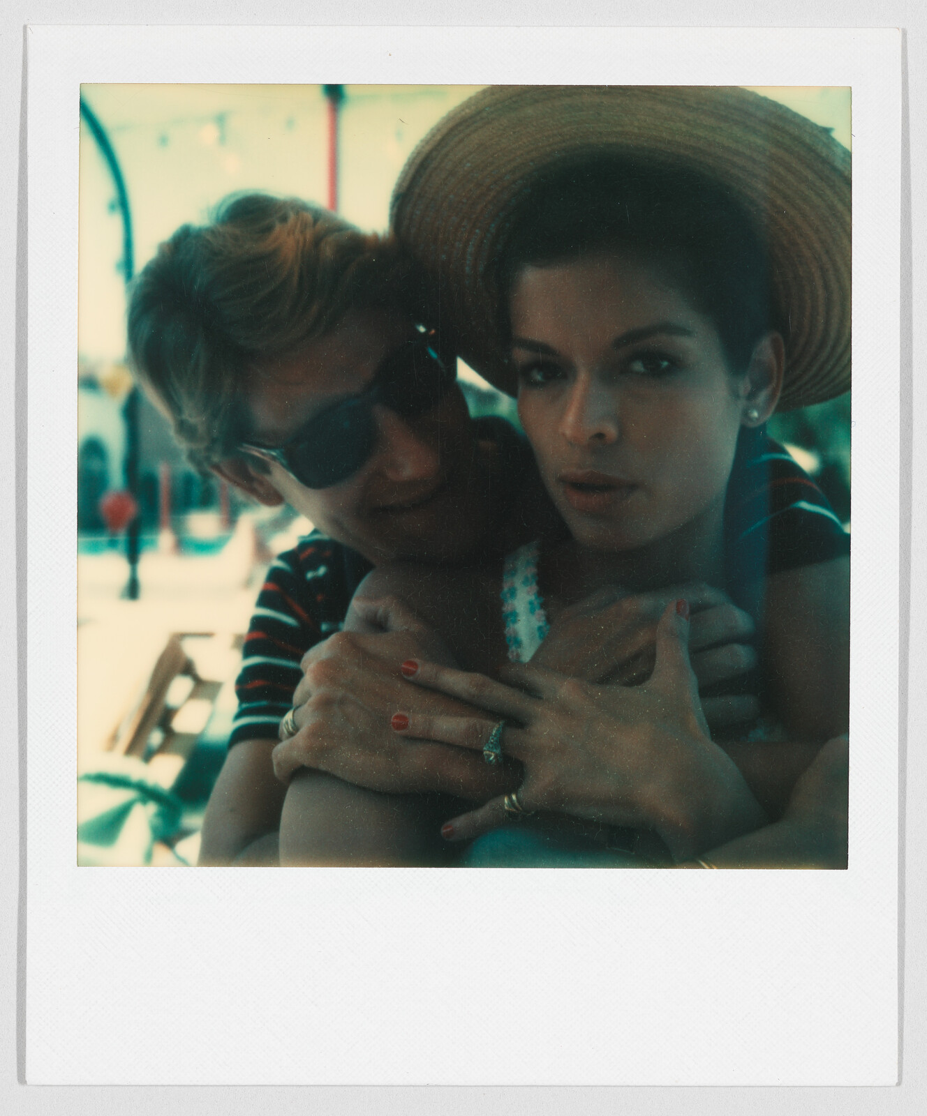 Two people sit close together; one wears sunglasses and hugs the other, who is wearing a wide-brimmed hat.