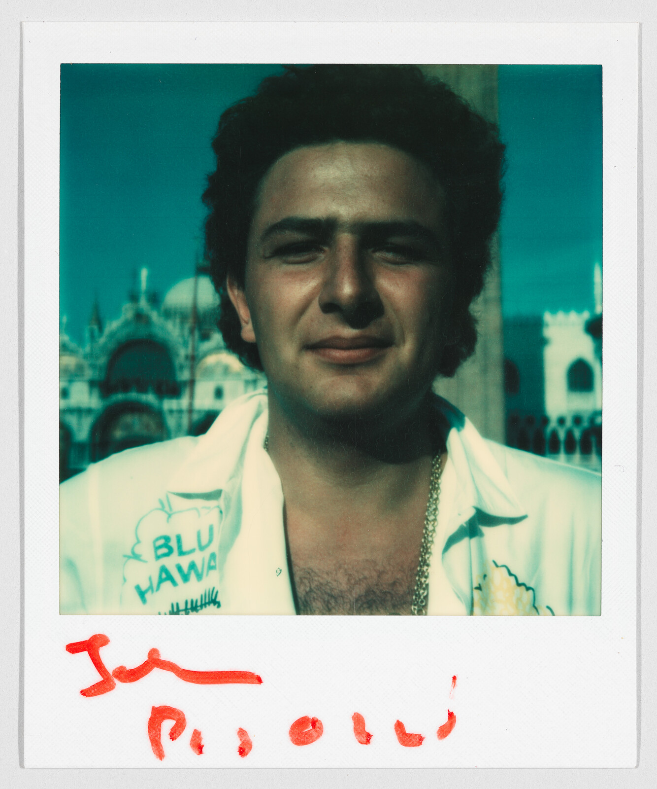 A man with curly hair wearing a white shirt that says "BLU HAWA" stands outdoors, smiling slightly.