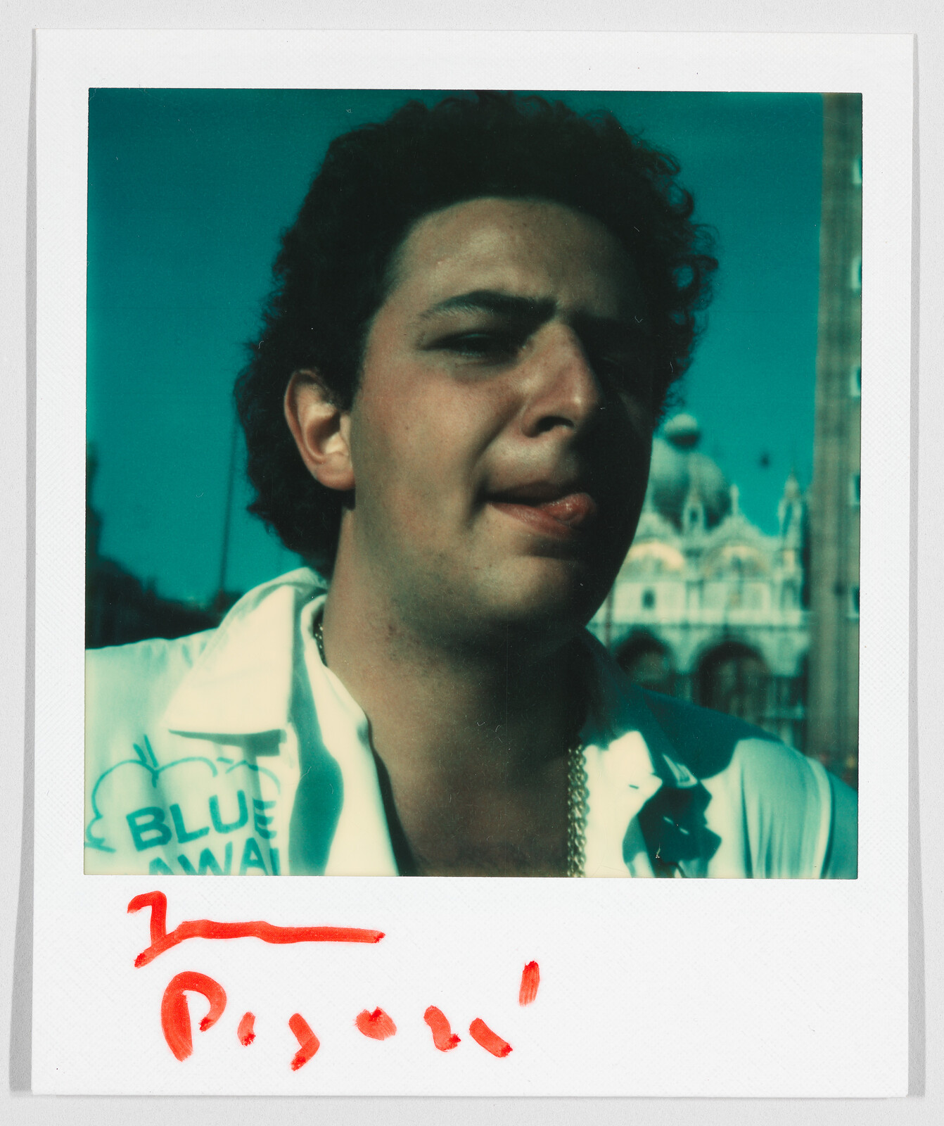 A man with curly hair sticks out his tongue, wearing a shirt with "BLUE" visible and a gold chain.