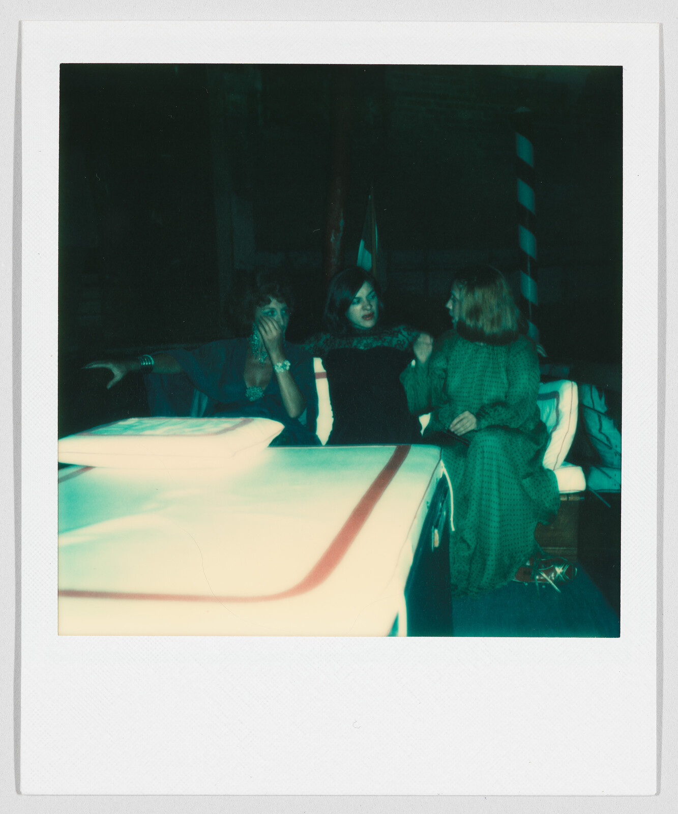 Three people sit together on a bench, talking and laughing, with one person covering their mouth.