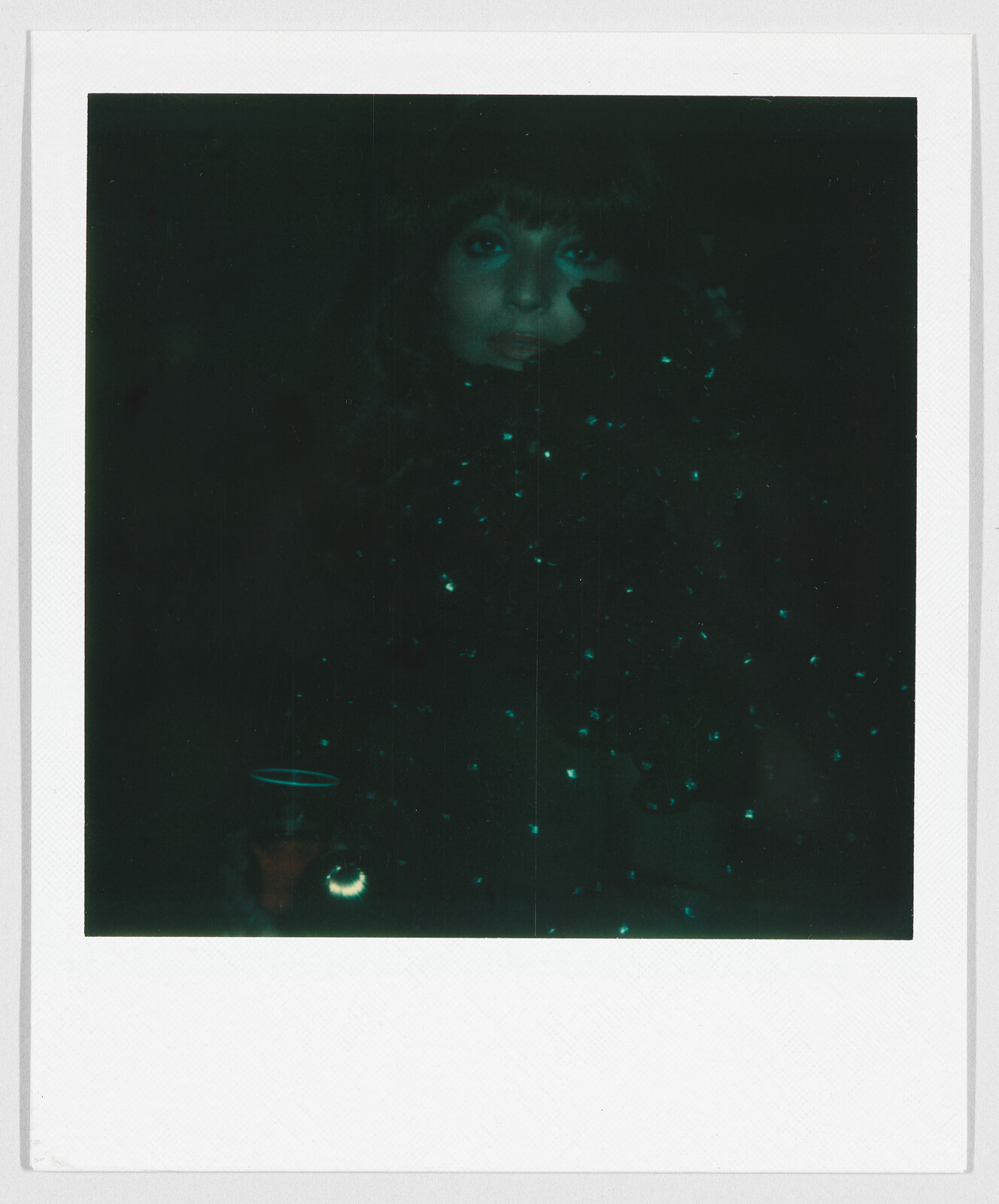 A person with long hair looks toward the camera in a dark setting with small glowing spots.