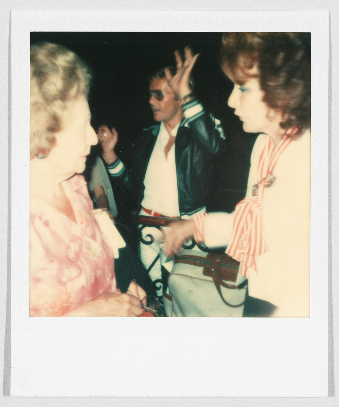 Three people are talking closely; one man in sunglasses gestures with his hands while two women face each other.