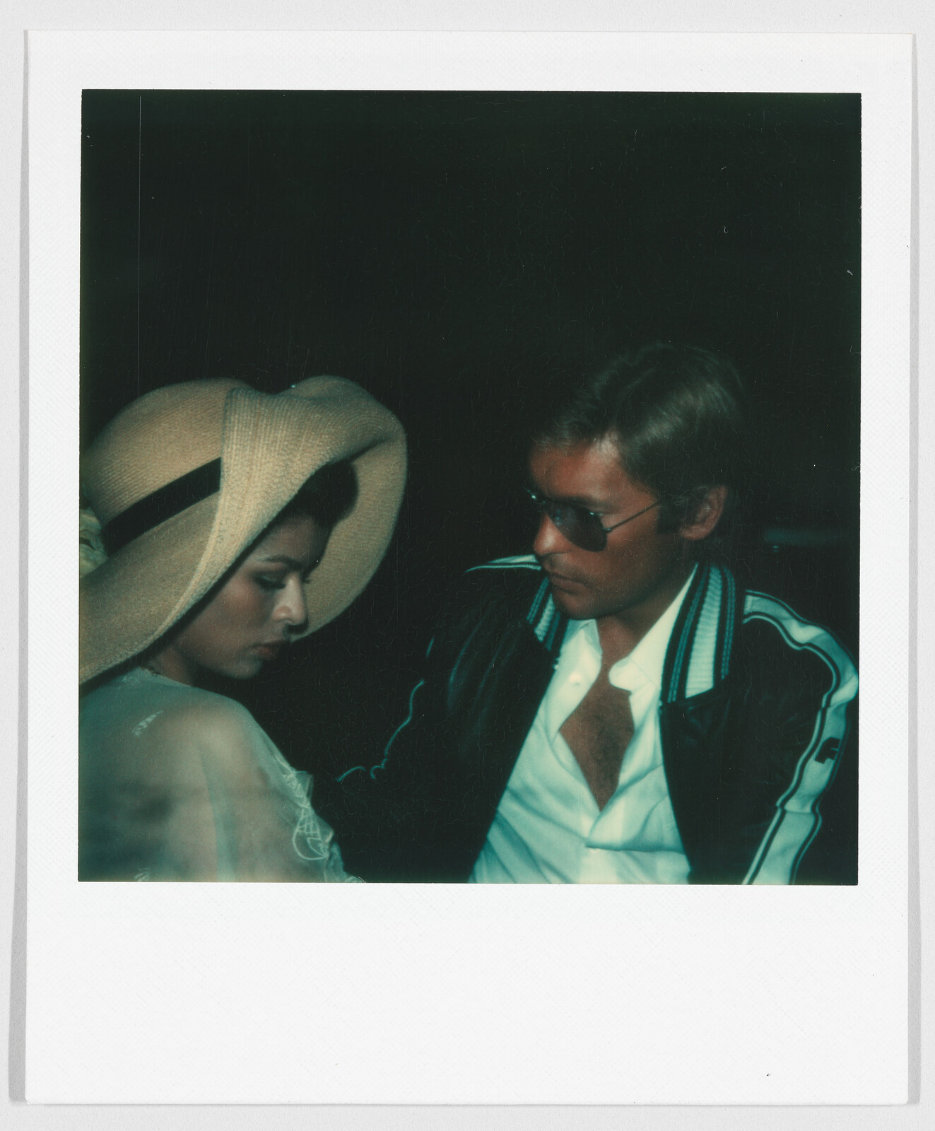 A woman in a wide-brimmed hat sits beside a man wearing sunglasses and a jacket, both looking down.