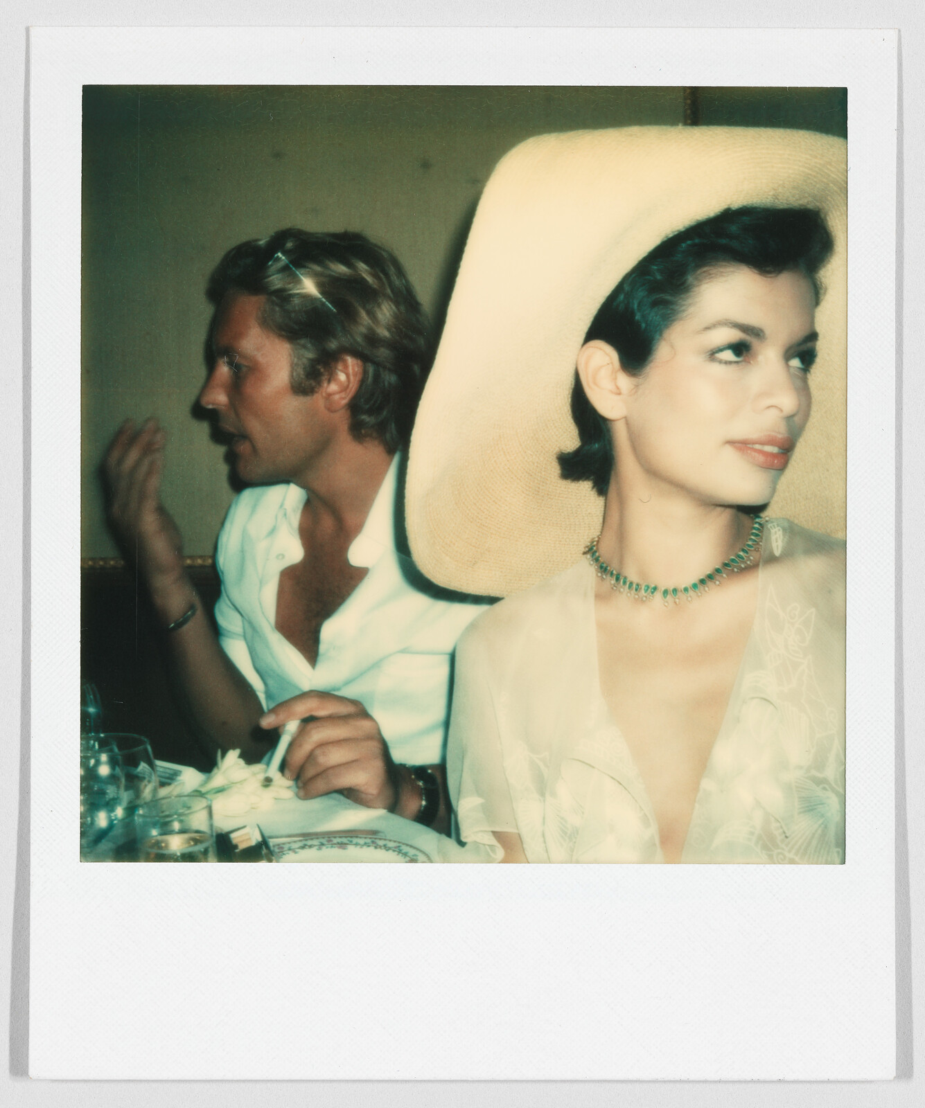A woman in a large hat sits at a table beside a man who is talking and holding a cigarette.