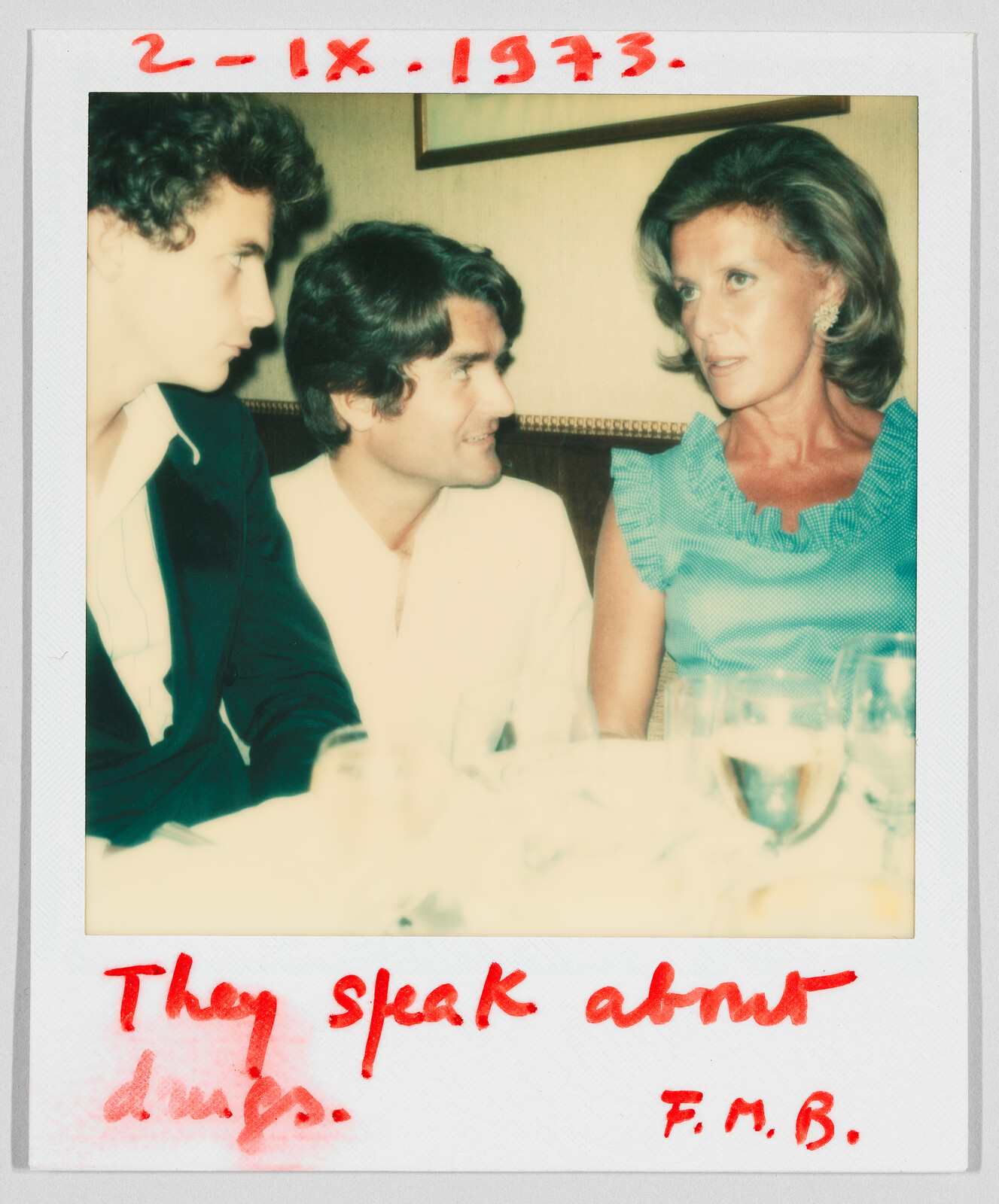 Three people sit at a table talking. Handwritten text says, “They speak about drugs. 2-IX-1973. F.M.B.”