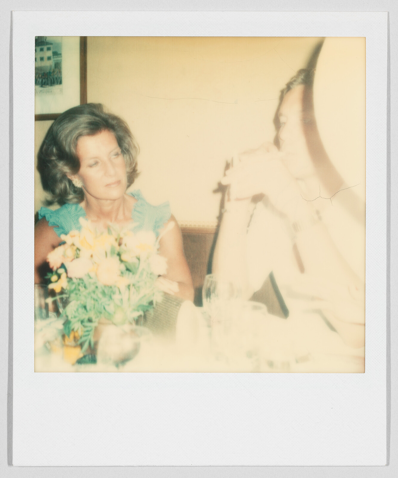 Two people sit at a table with flowers and drinks; one person looks at the other, who is blurred.