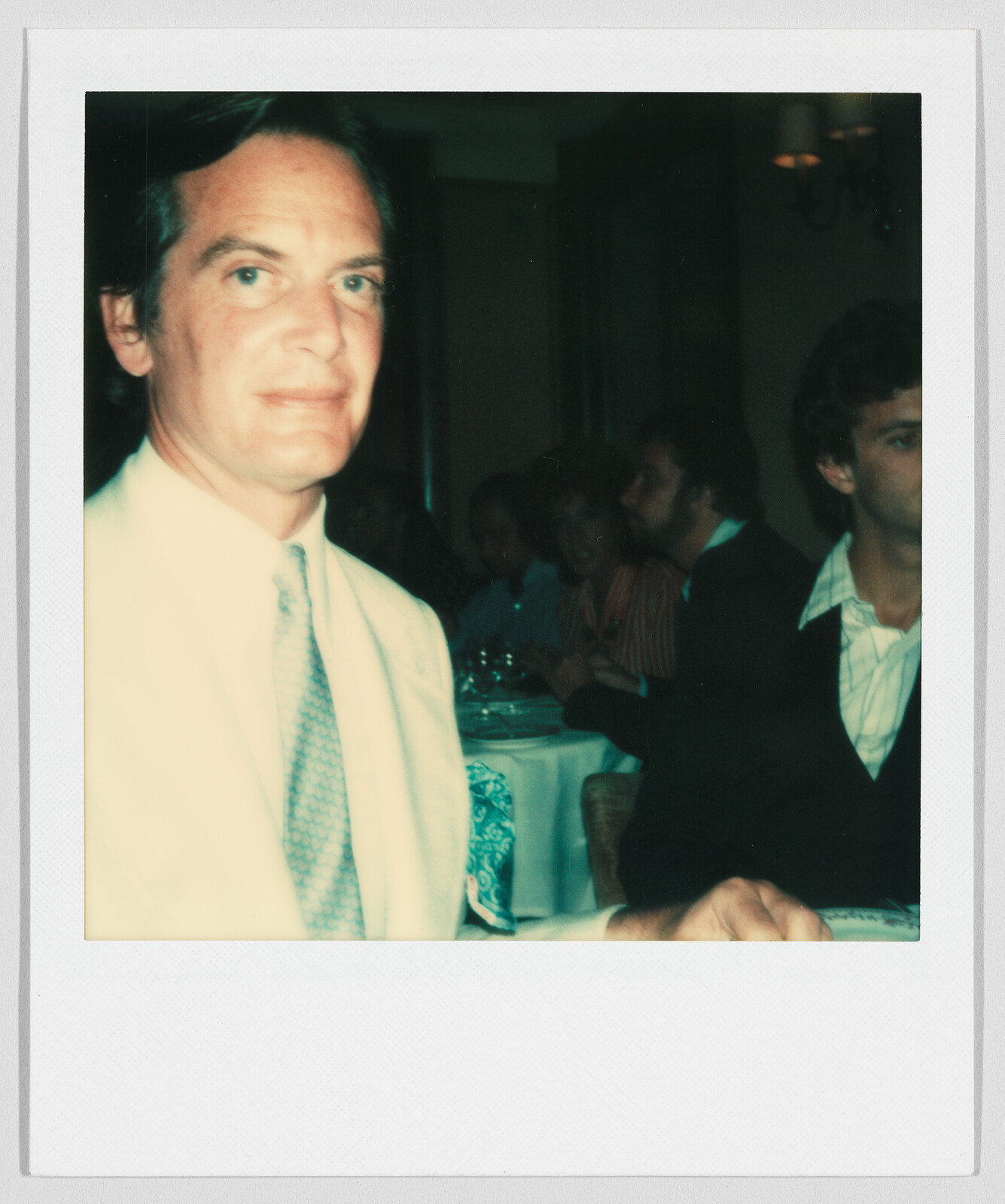 Man in a light suit and tie sits at a table in a restaurant with other people in the background.