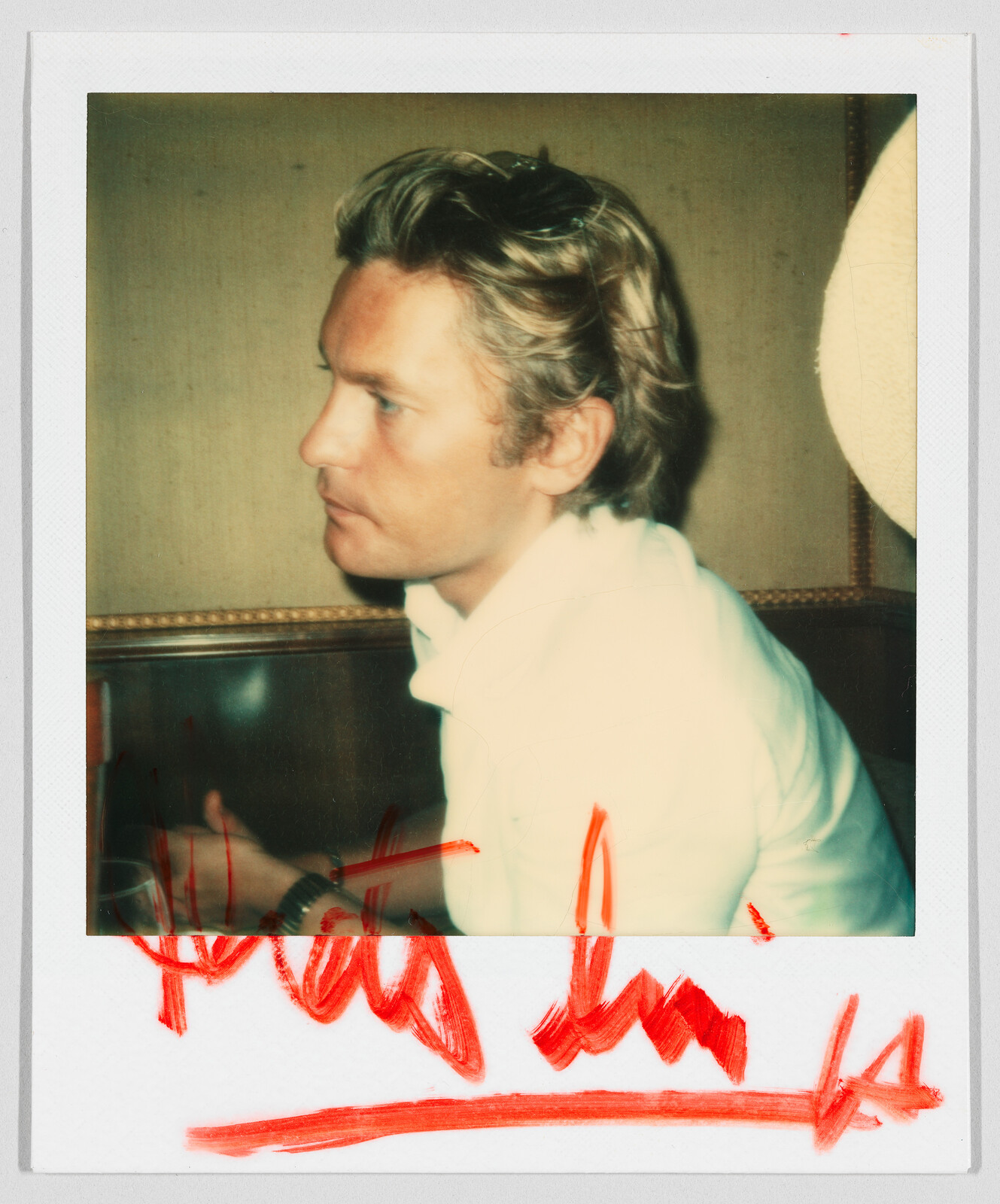 A man with blond hair sits sideways, wearing a white shirt. Red handwriting is visible at the bottom.