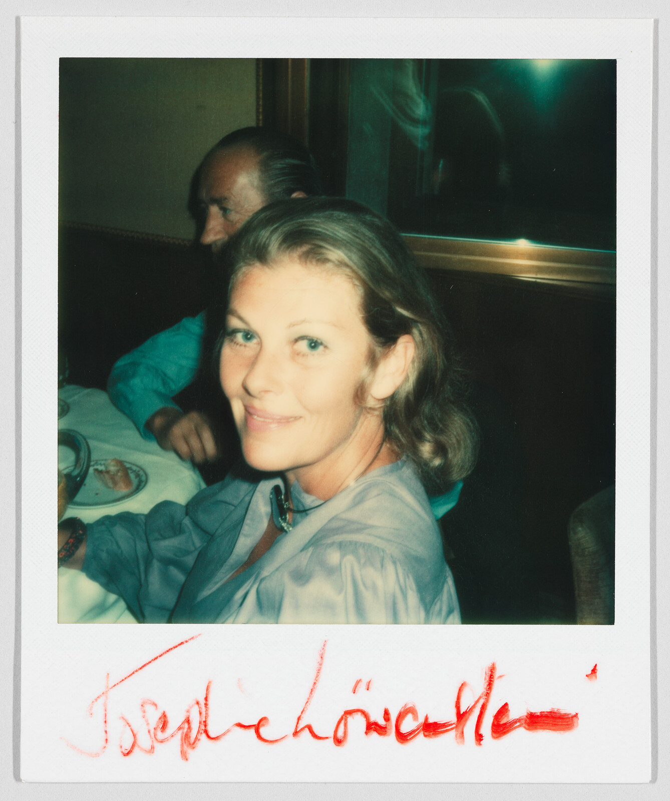 Woman with light brown hair smiles at a table in a restaurant; red signature is written below.