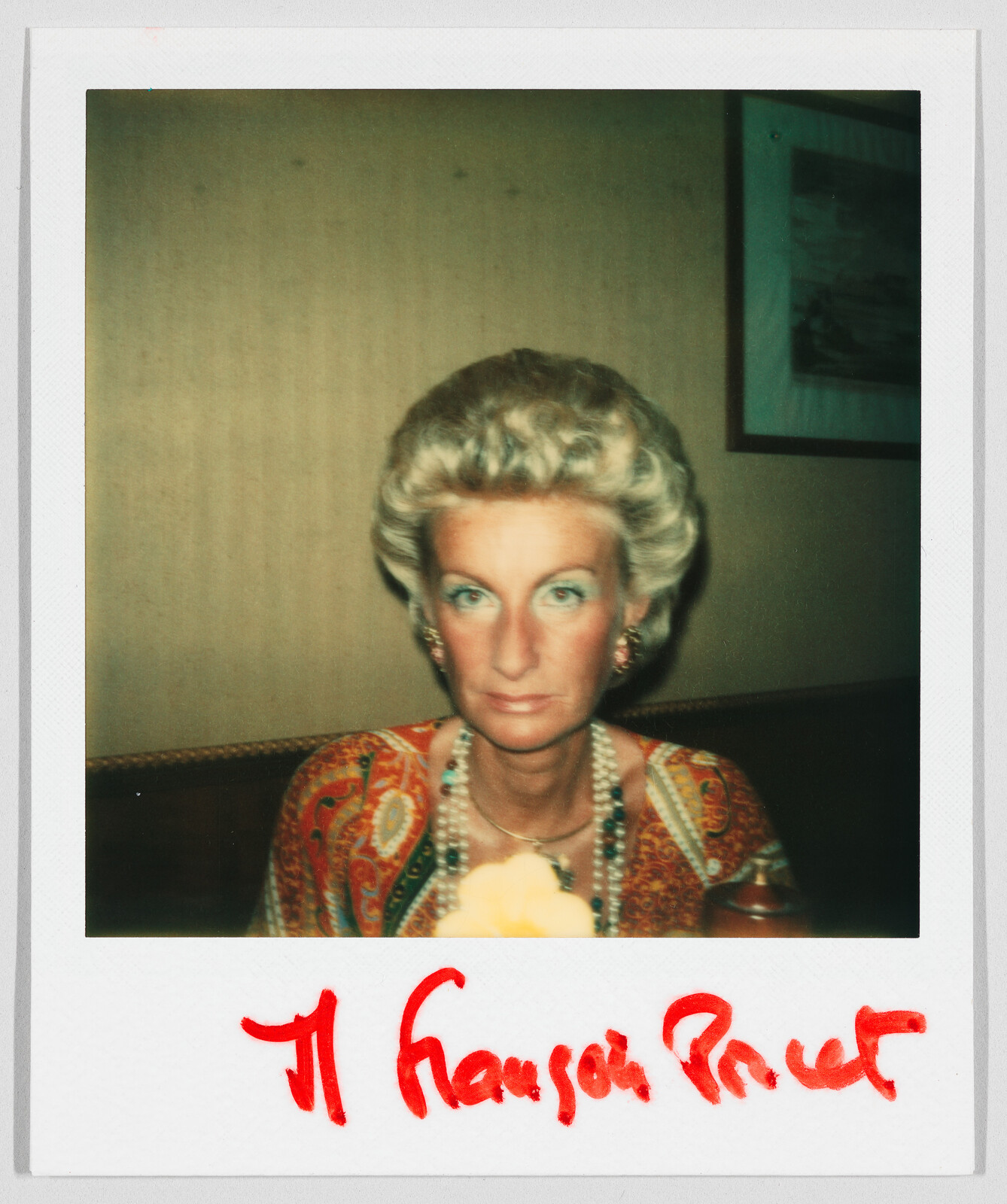 Woman with styled blonde hair and colorful clothing sits at a table; red handwriting appears at the bottom.