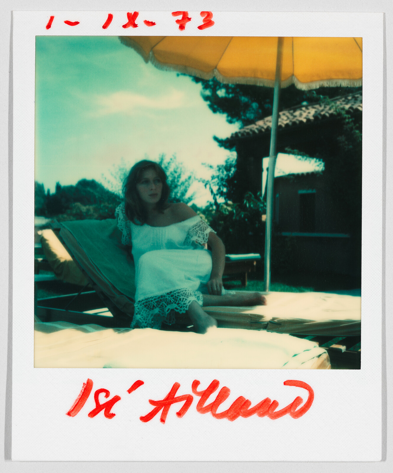 A woman in a white dress sits on a lounge chair under a large yellow umbrella outdoors. Handwritten text reads "1-IX-73" and "Isi Allard."