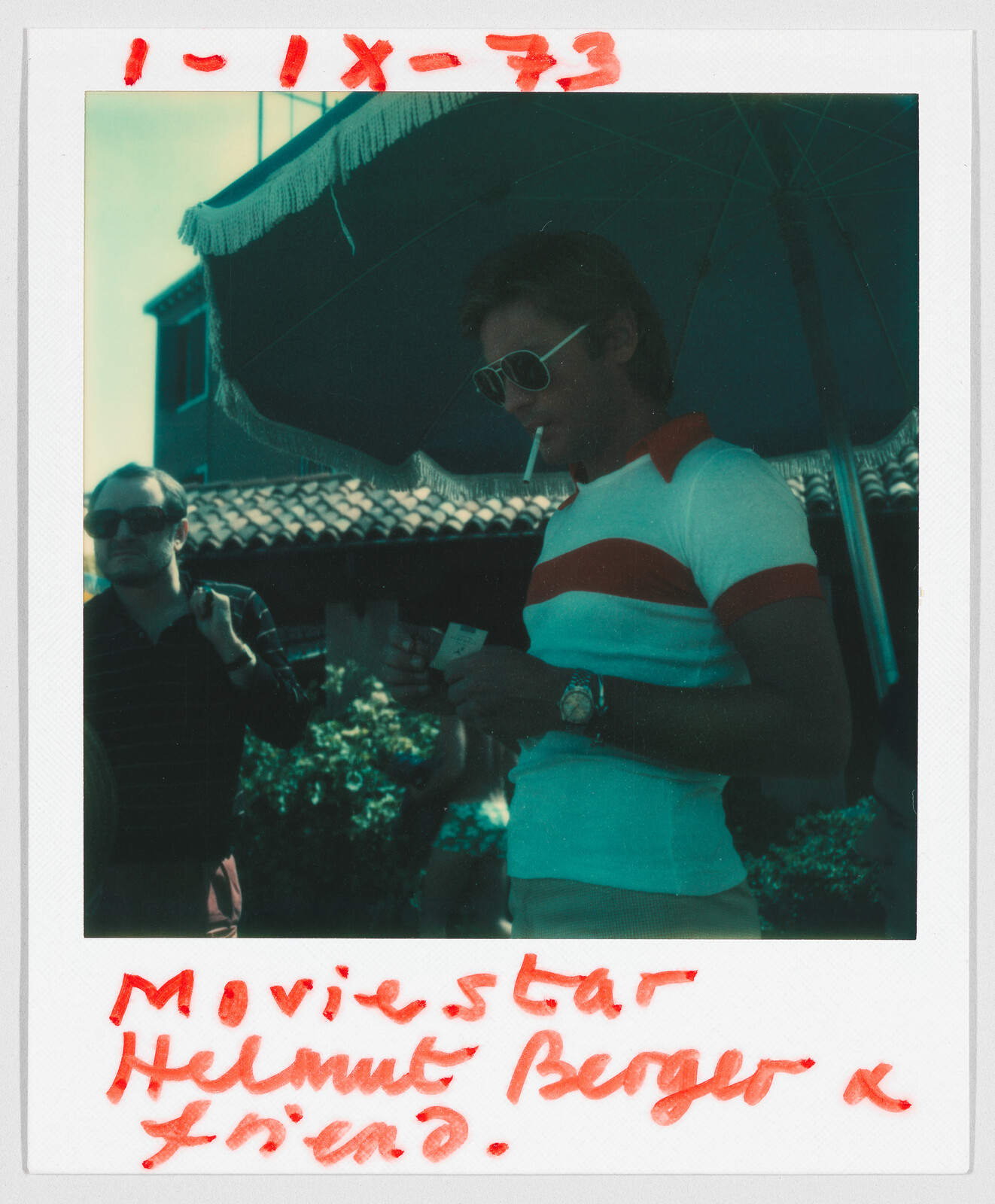 Two men stand outdoors under a large umbrella; handwritten text reads “Moviestar Helmut Berger & friend.”