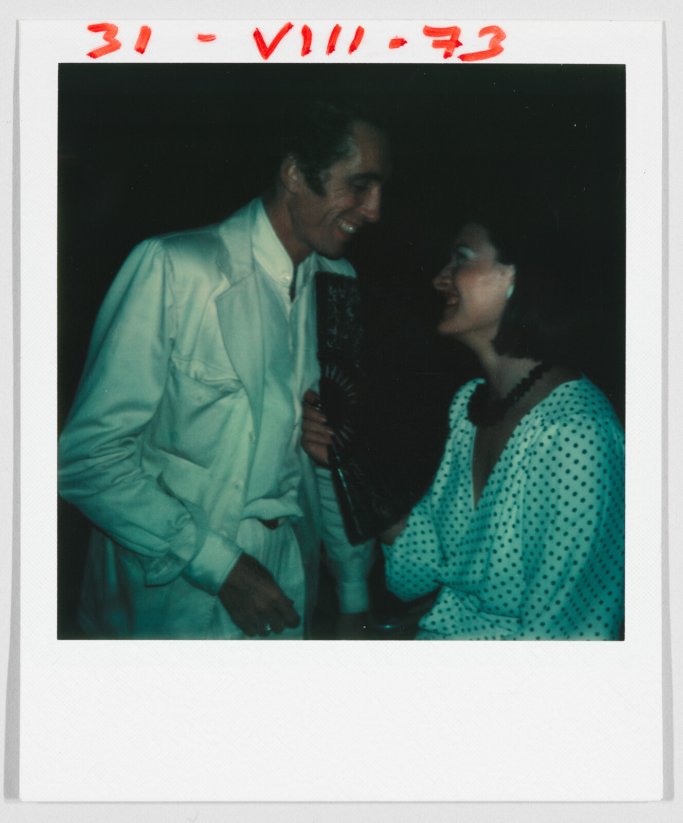 A man in a white suit and a woman in a polka dot dress smile and talk together. The date "31-VIII-73" is written in red at the top.