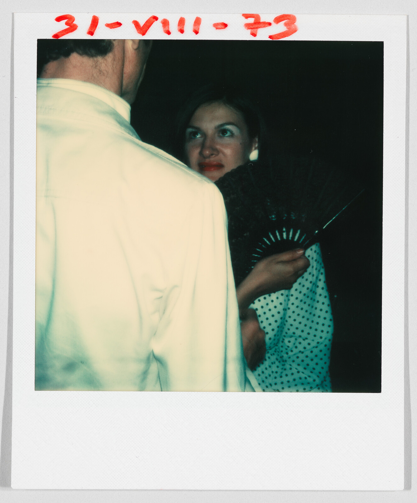 A woman in a polka dot dress holds a fan, looking at a man in a white jacket. Date “31-VIII-73” is written in red at the top.