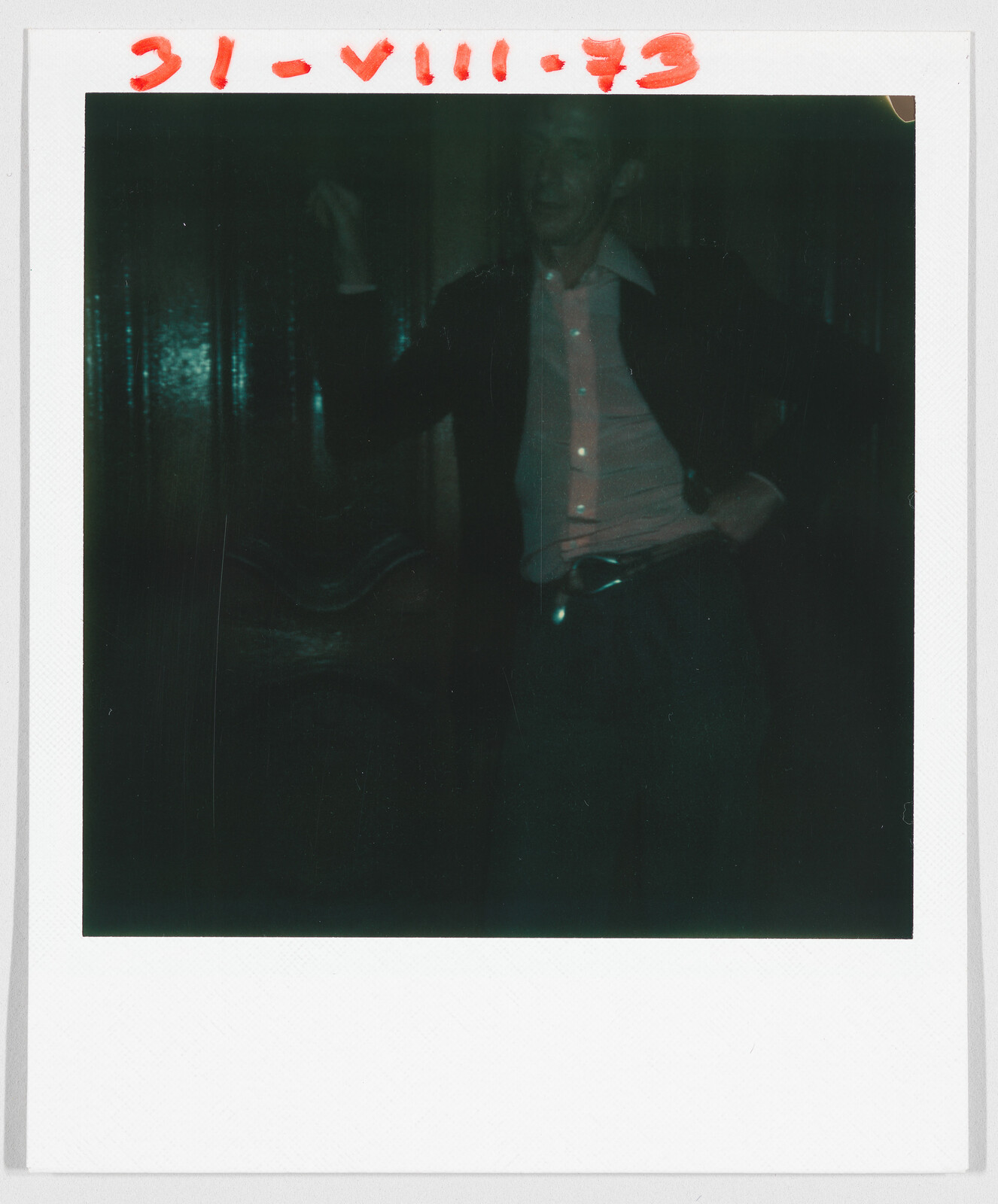 A person stands in a dimly lit room, wearing a light shirt and dark jacket. Date: 31-VIII-73.