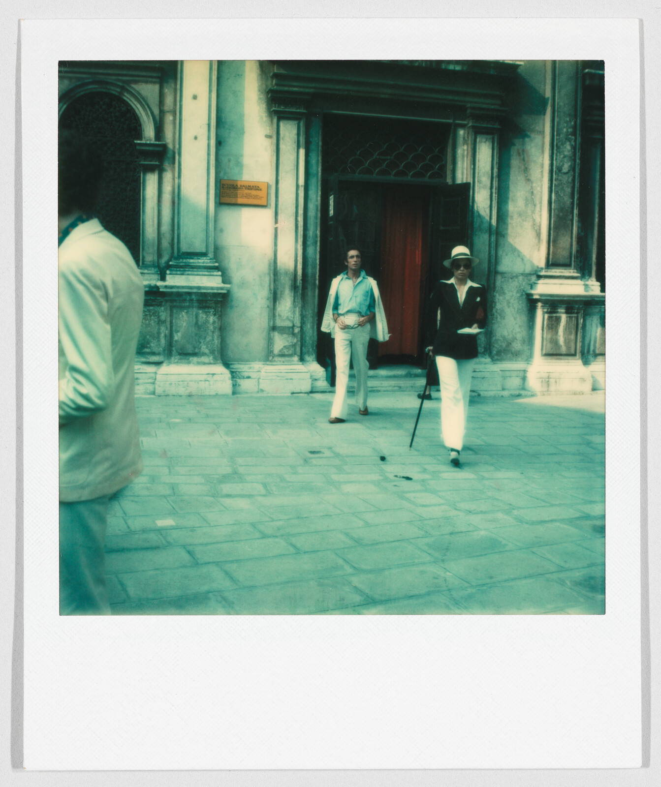 Two people in light clothing stand and walk outside a building with a yellow sign and open door.