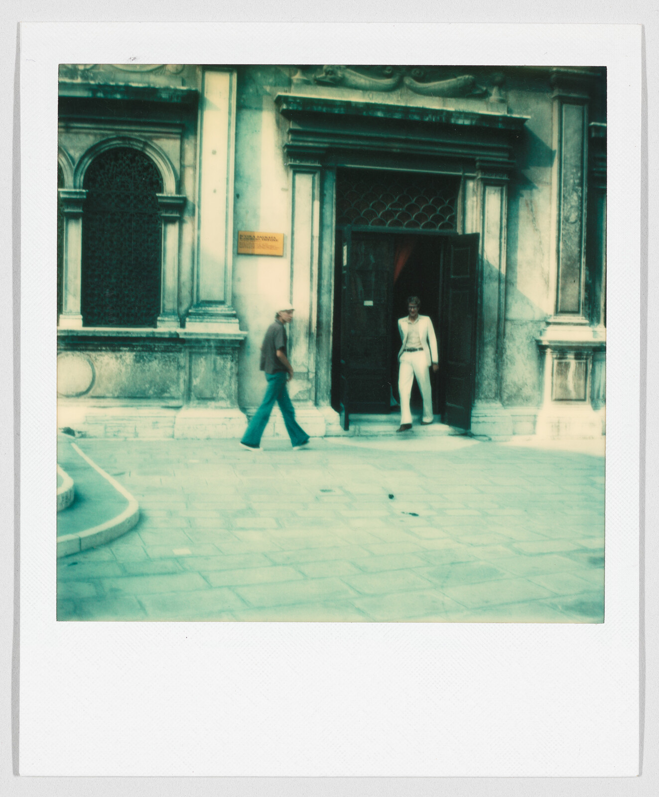 A person in white stands in a doorway while another person walks past on a stone street.