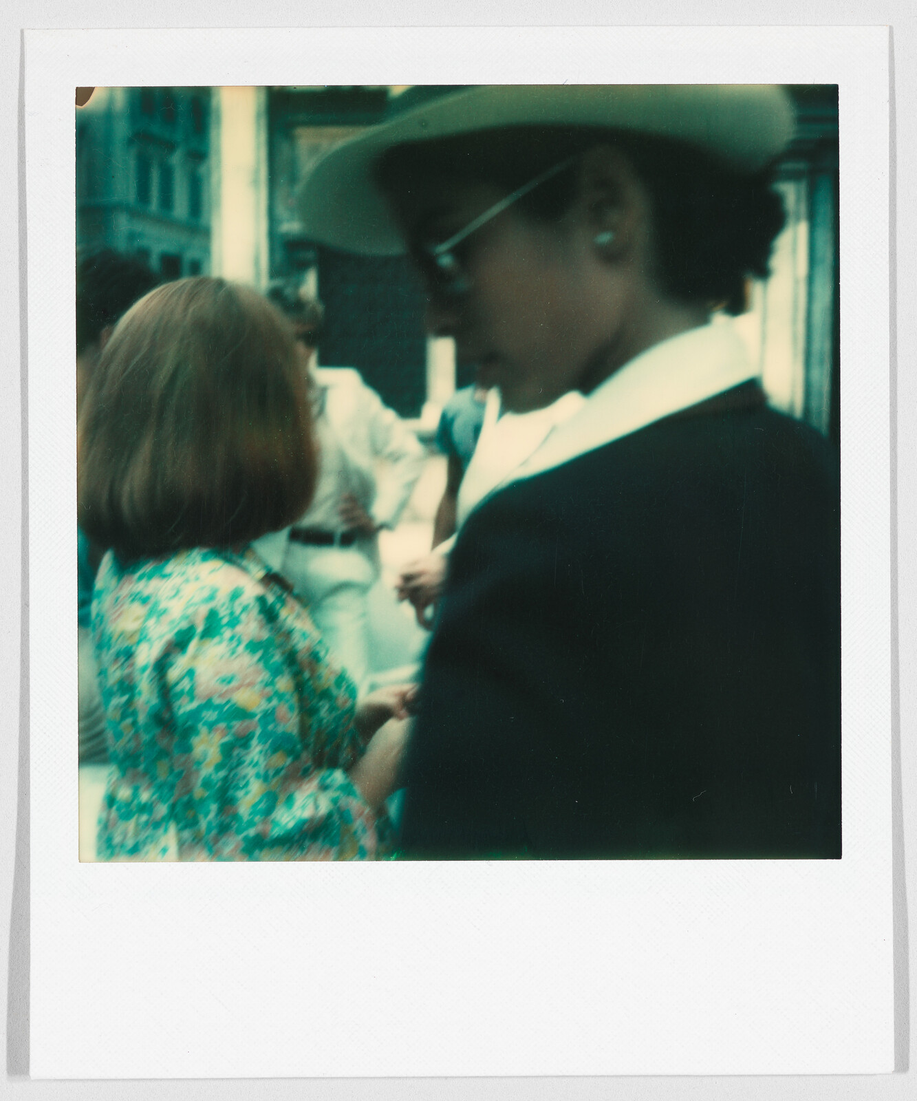 Several people stand close together outdoors; one person wears a hat and sunglasses, another wears a floral shirt.