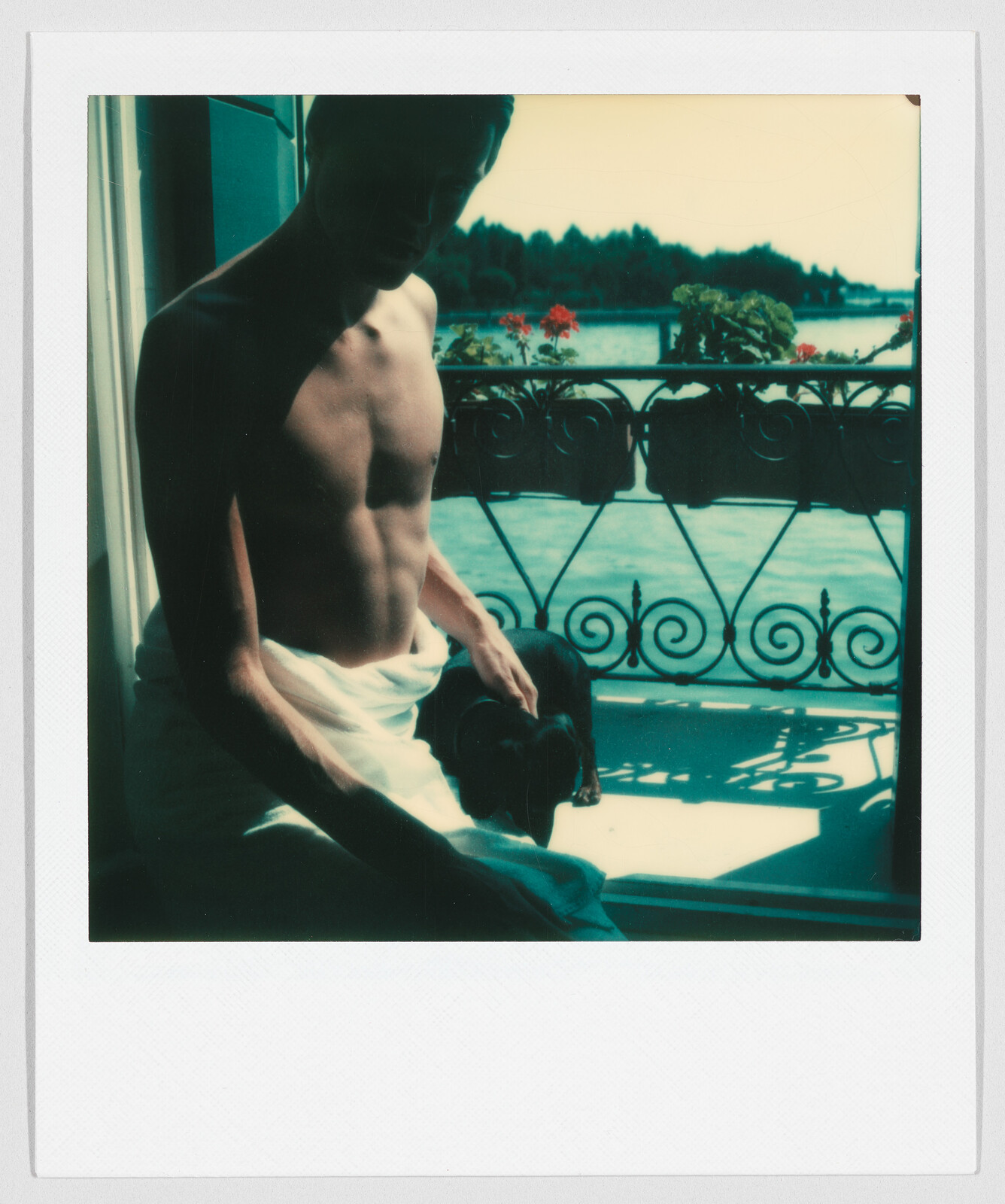 Shirtless person with a towel sits by a balcony, with a lake and trees in the background.