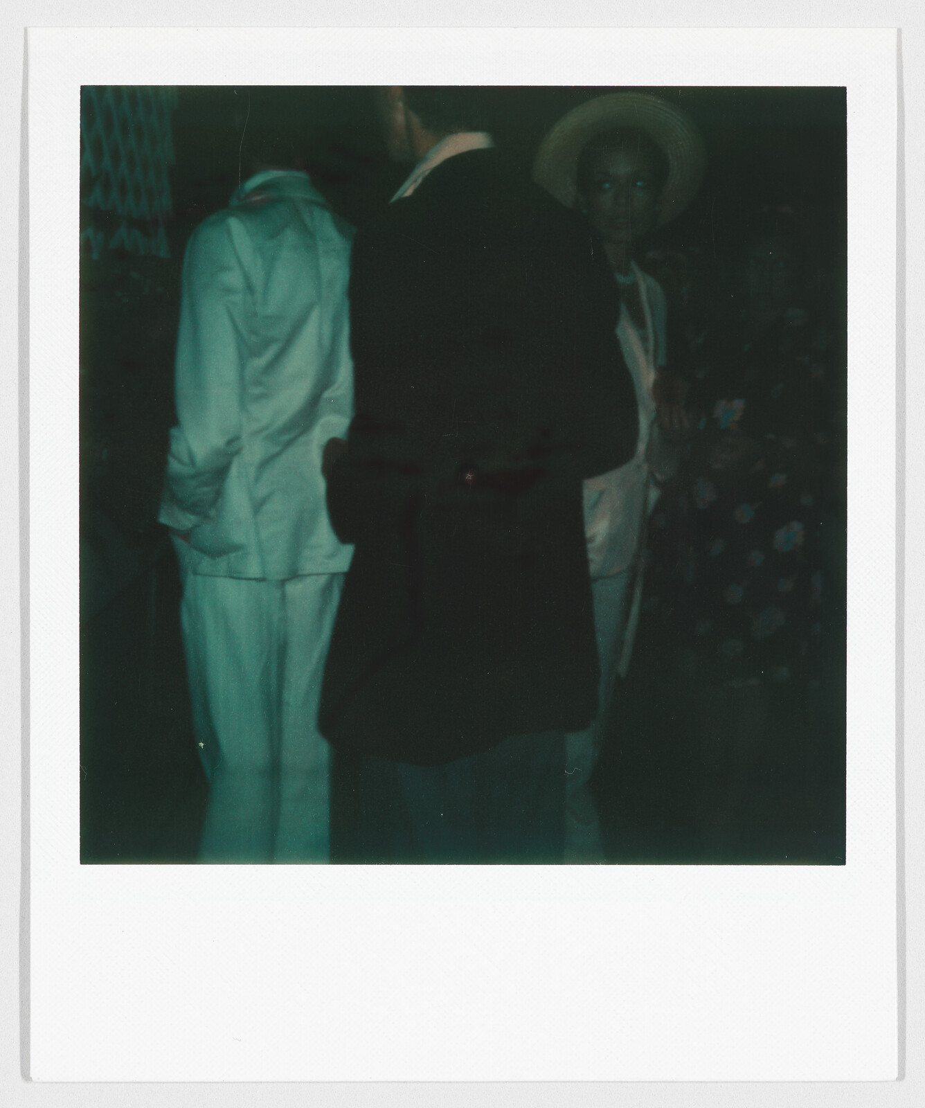 Three people stand together in dim lighting; one wears a light suit and another wears a hat.