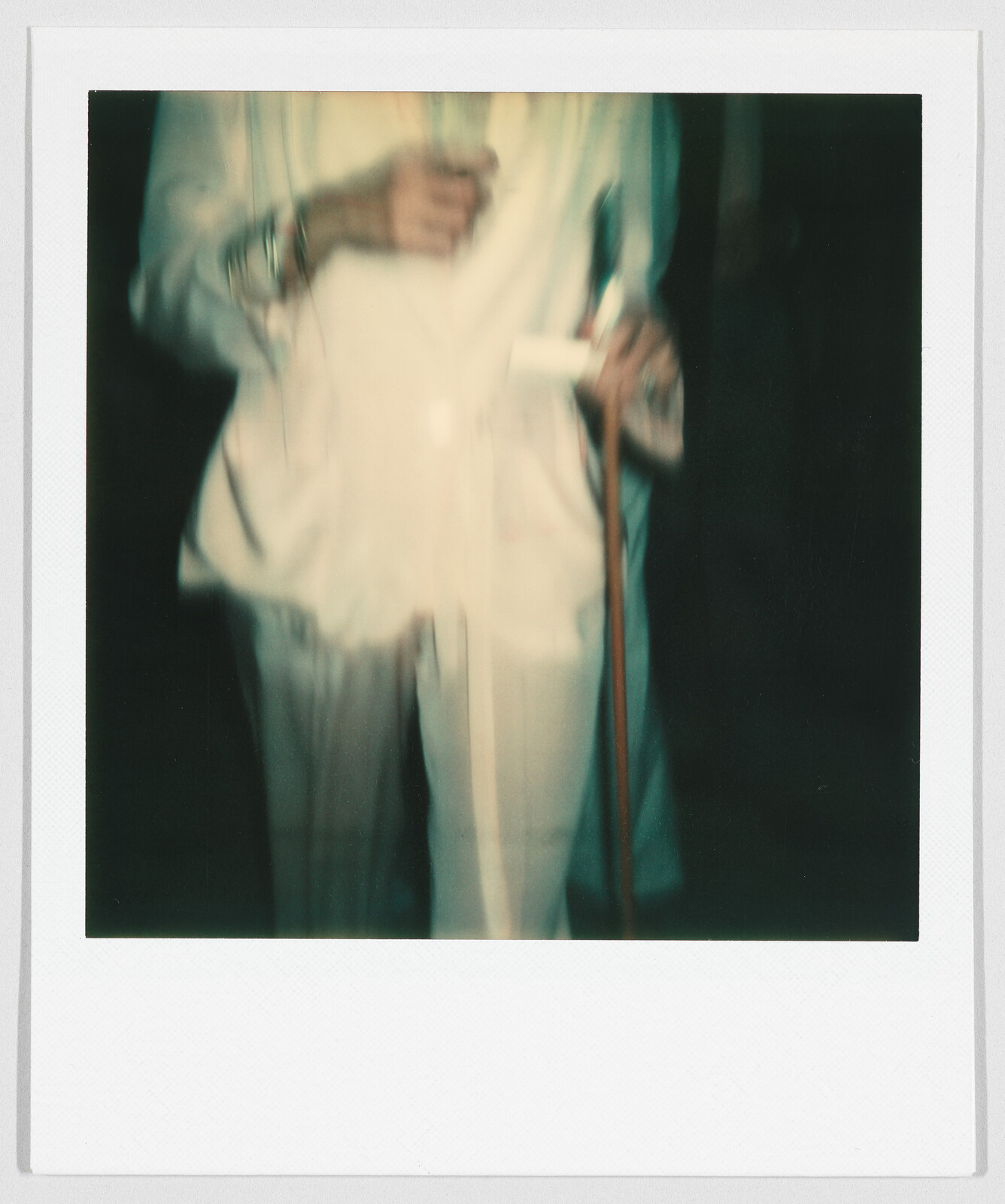 Person in a light-colored suit holding a cane and a drink, image is blurry and unclear.