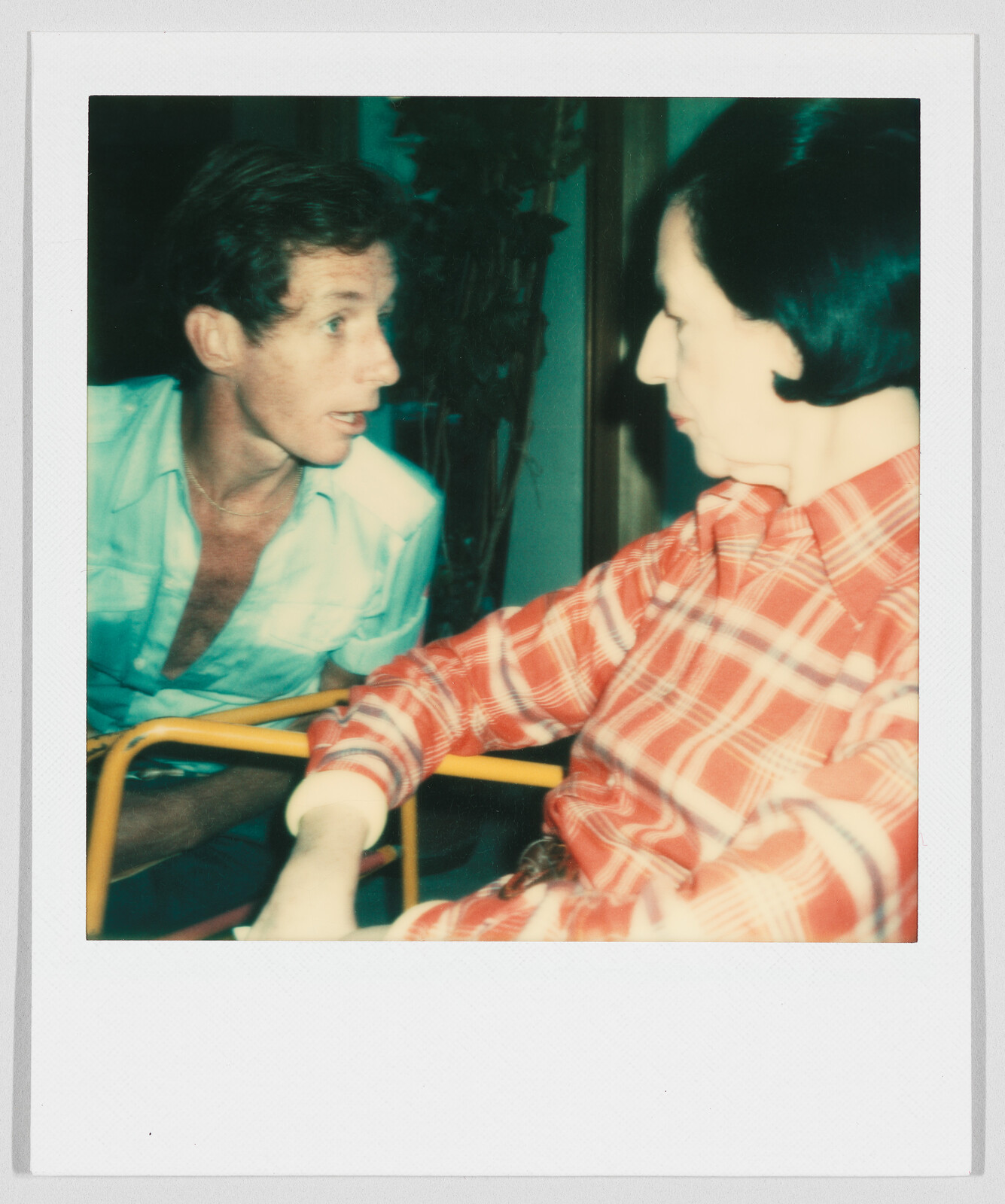 Two people sit close together, talking. One wears a red plaid shirt, the other a light blue shirt.