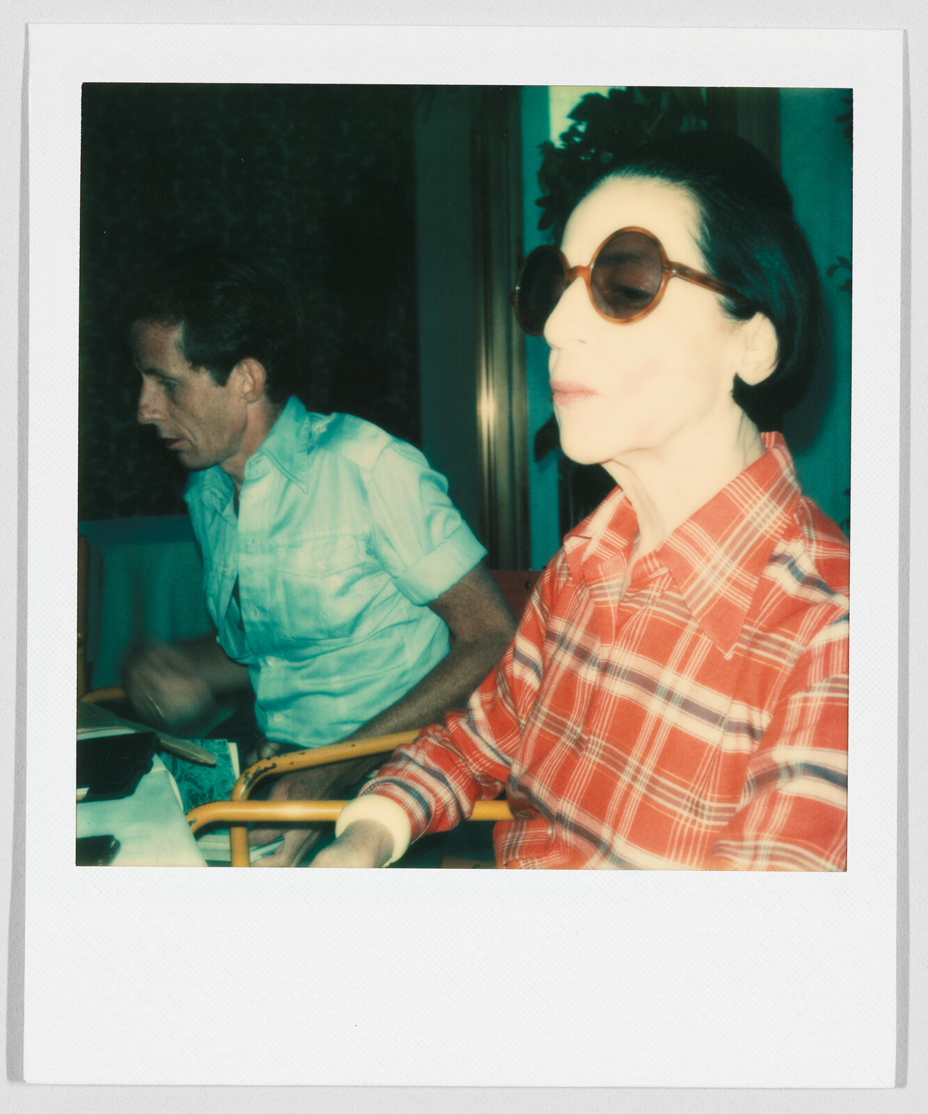 Two people sit at a table; one wears large sunglasses and a red plaid shirt, the other looks down.