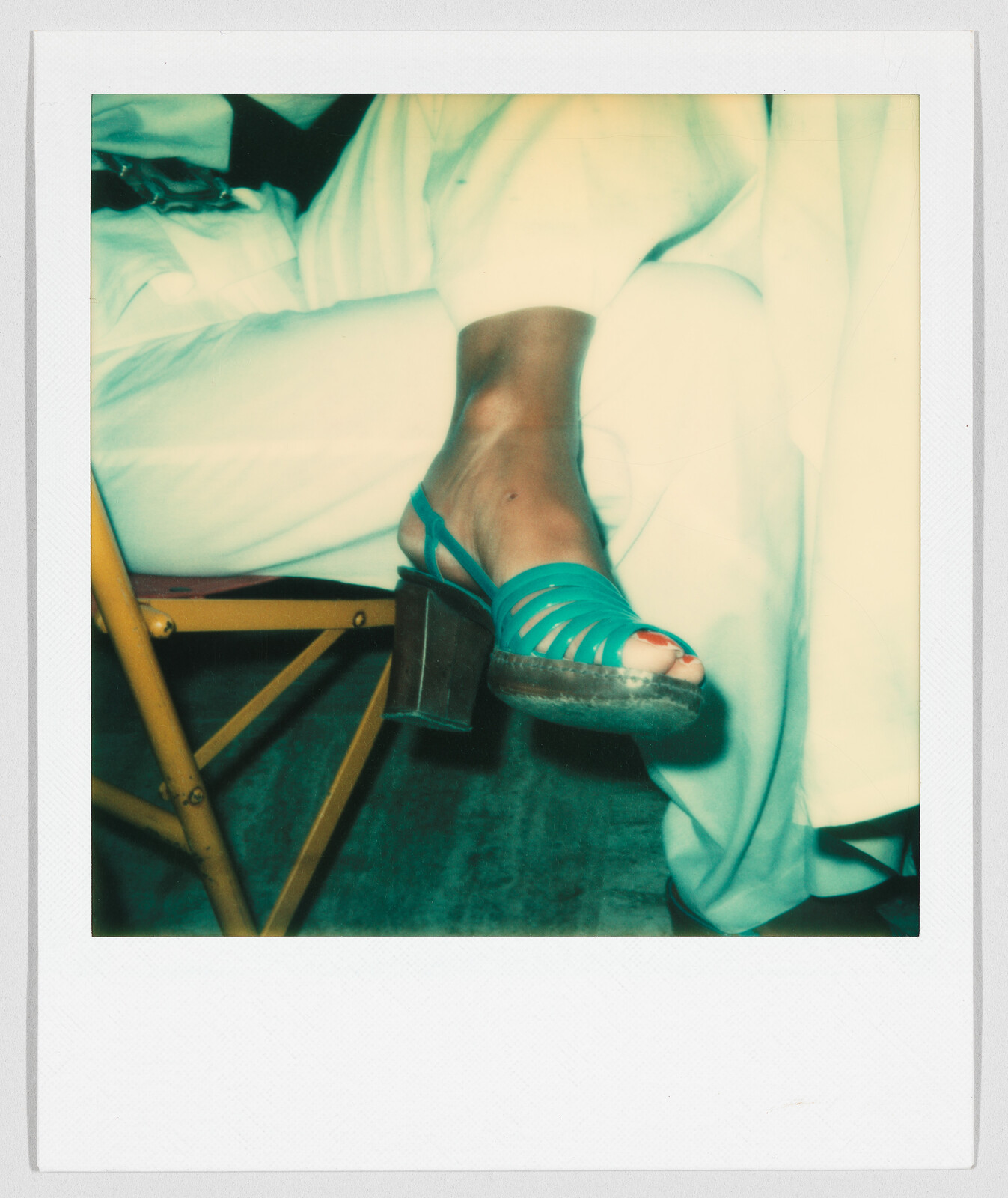 A person wearing white pants sits with legs crossed, showing a turquoise sandal with a chunky heel.