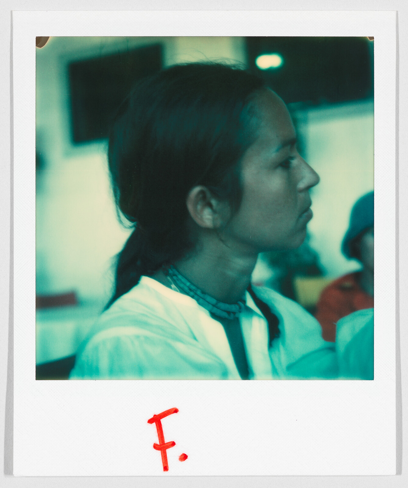 A person with long hair tied back looks to the side; the letter "F." is written in red below.