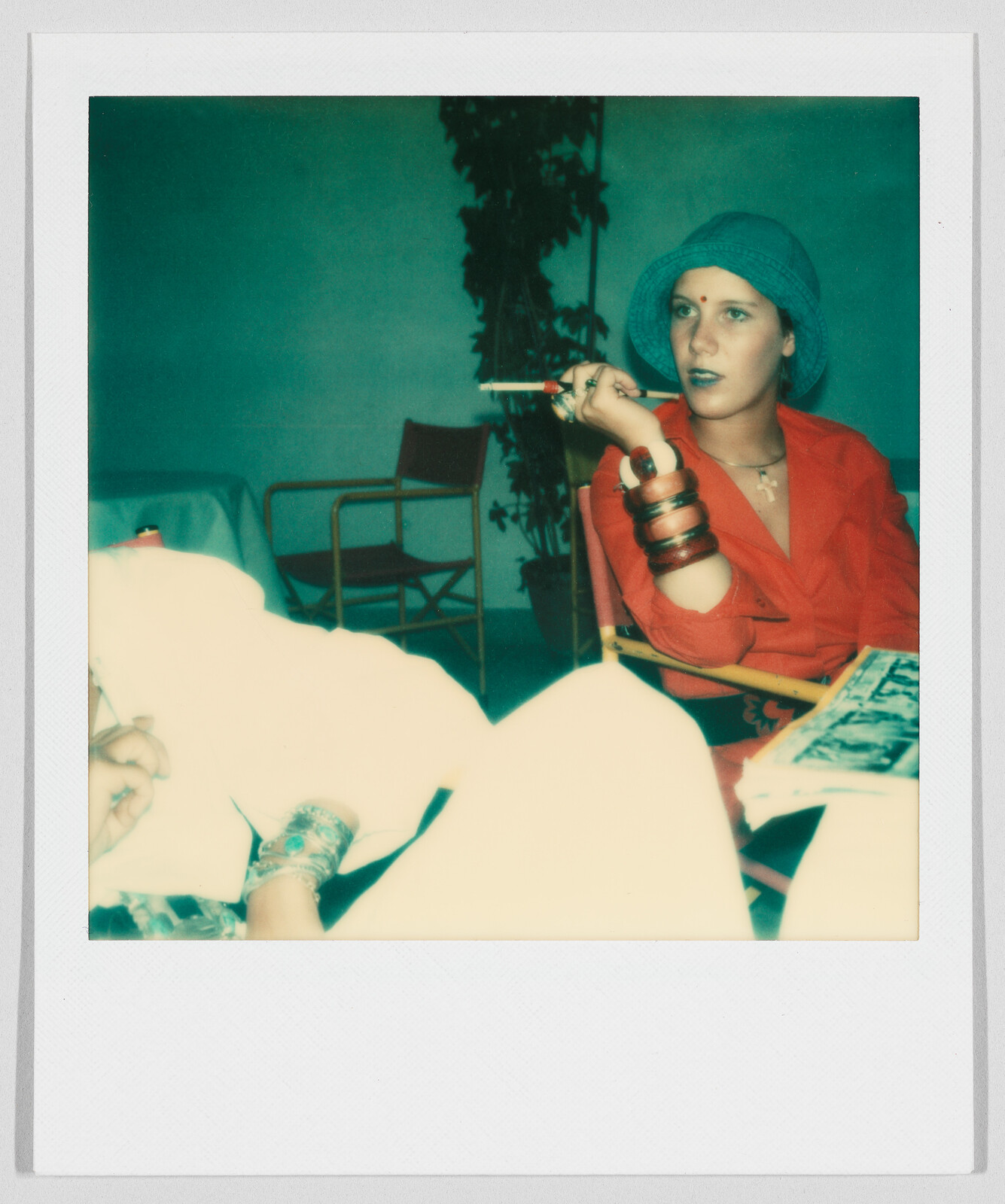 A woman in a blue hat and red shirt holds a cigarette holder, sitting at a table with another person.