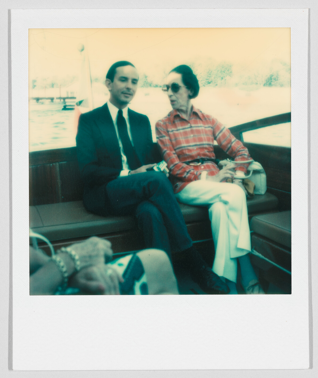 A man in a suit and a woman in a red plaid shirt sit together on a boat bench.