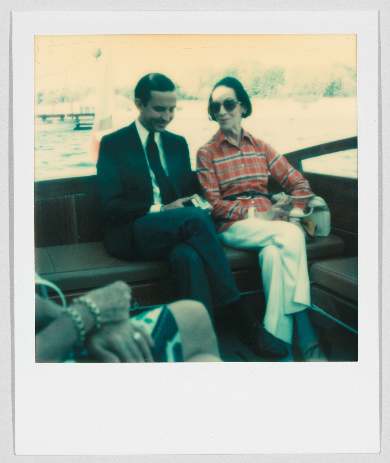 A man in a suit and a woman in sunglasses sit together on a boat, talking and smiling.