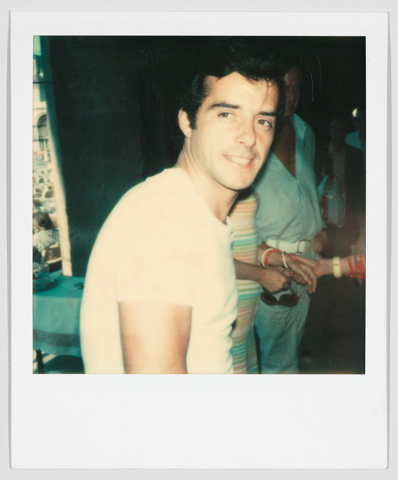 A man in a white t-shirt smiles at the camera indoors, with people and a table in the background.