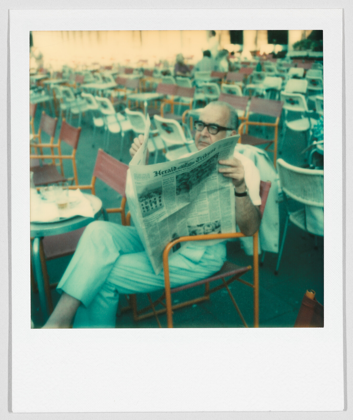 Man sitting at an outdoor café, reading a newspaper with the headline “Herald Tribune” visible.