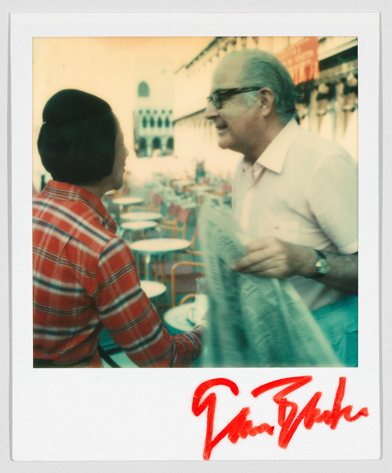 Two people greet each other outdoors at a café with empty tables; one holds a newspaper. Red signature below.