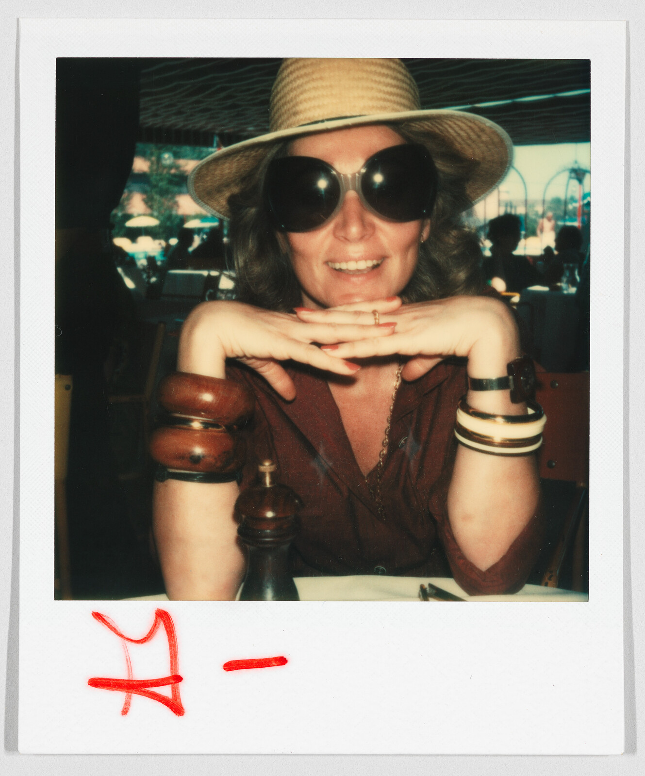Woman wearing a straw hat and large sunglasses, smiling with hands under chin at a restaurant table.