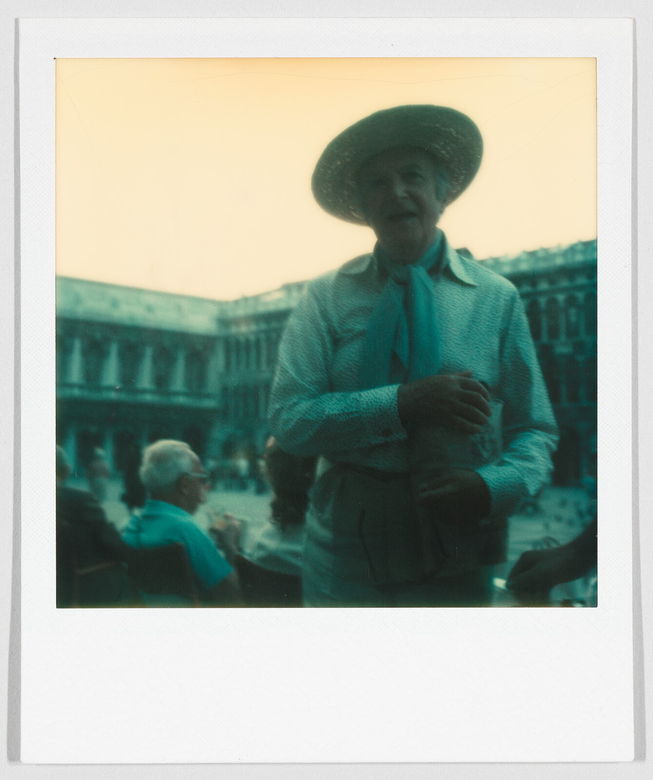 Person wearing a wide-brimmed hat and scarf stands outdoors, with people sitting and a large building behind.