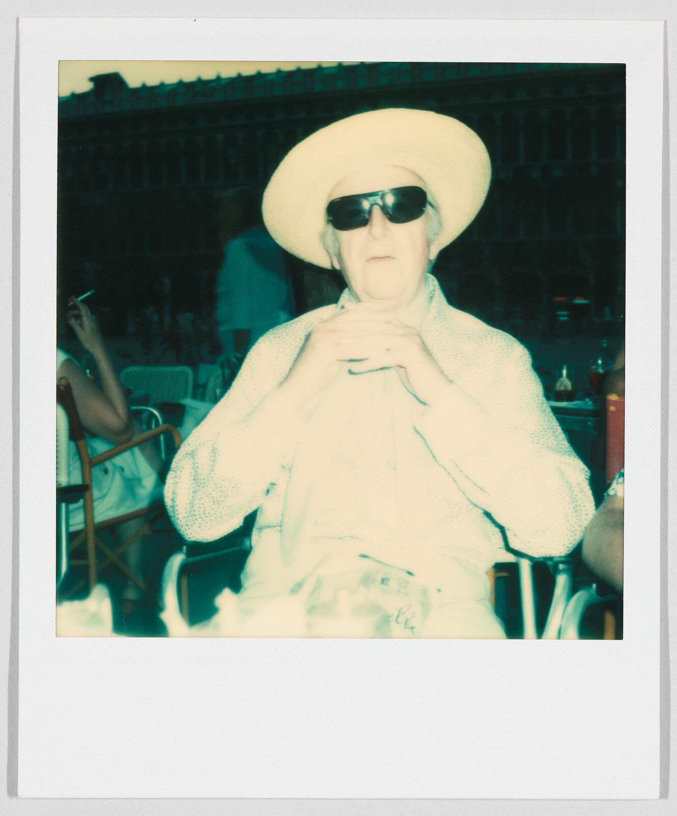 Older person wearing a wide-brimmed hat and dark sunglasses sits at an outdoor table with hands clasped.
