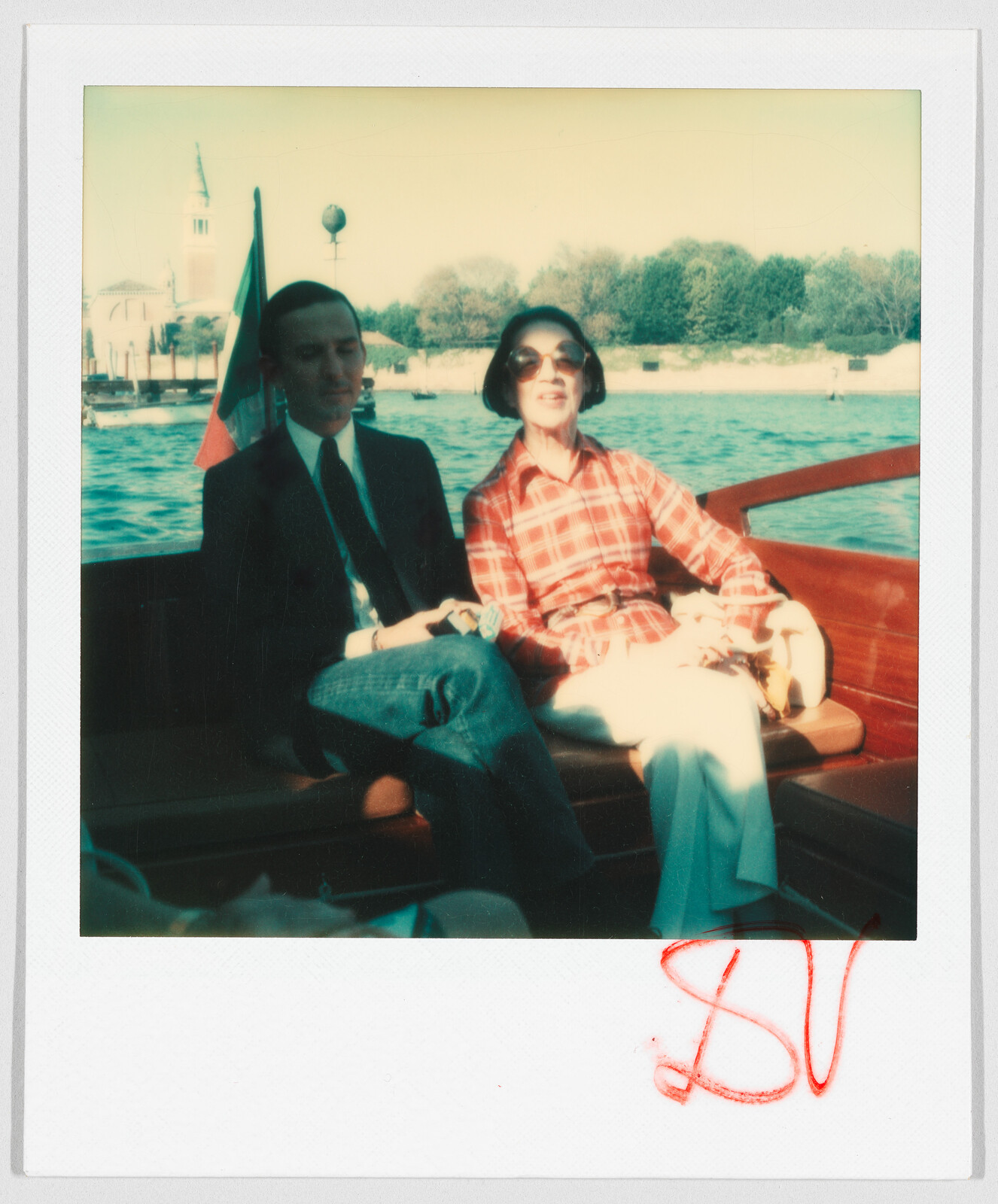 Two people sit together on a boat, one wearing a suit and the other in a red plaid shirt.