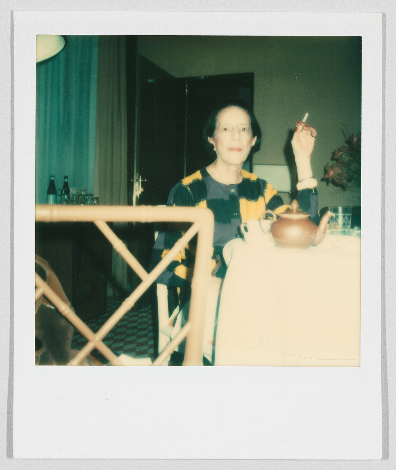 An older woman sits at a table holding a cigarette, with a teapot and cup in front of her.