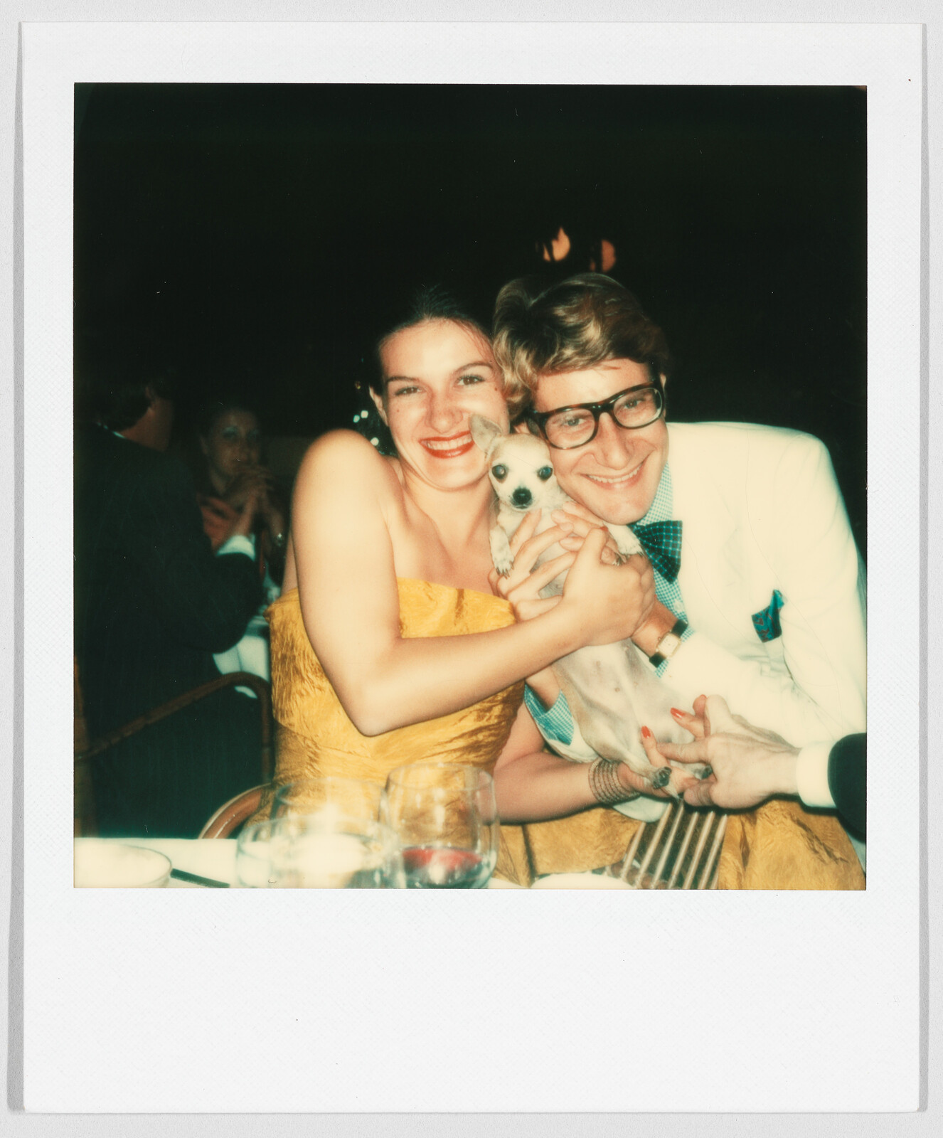 Smiling woman and man sitting close together, holding a small dog at a table with drinks.