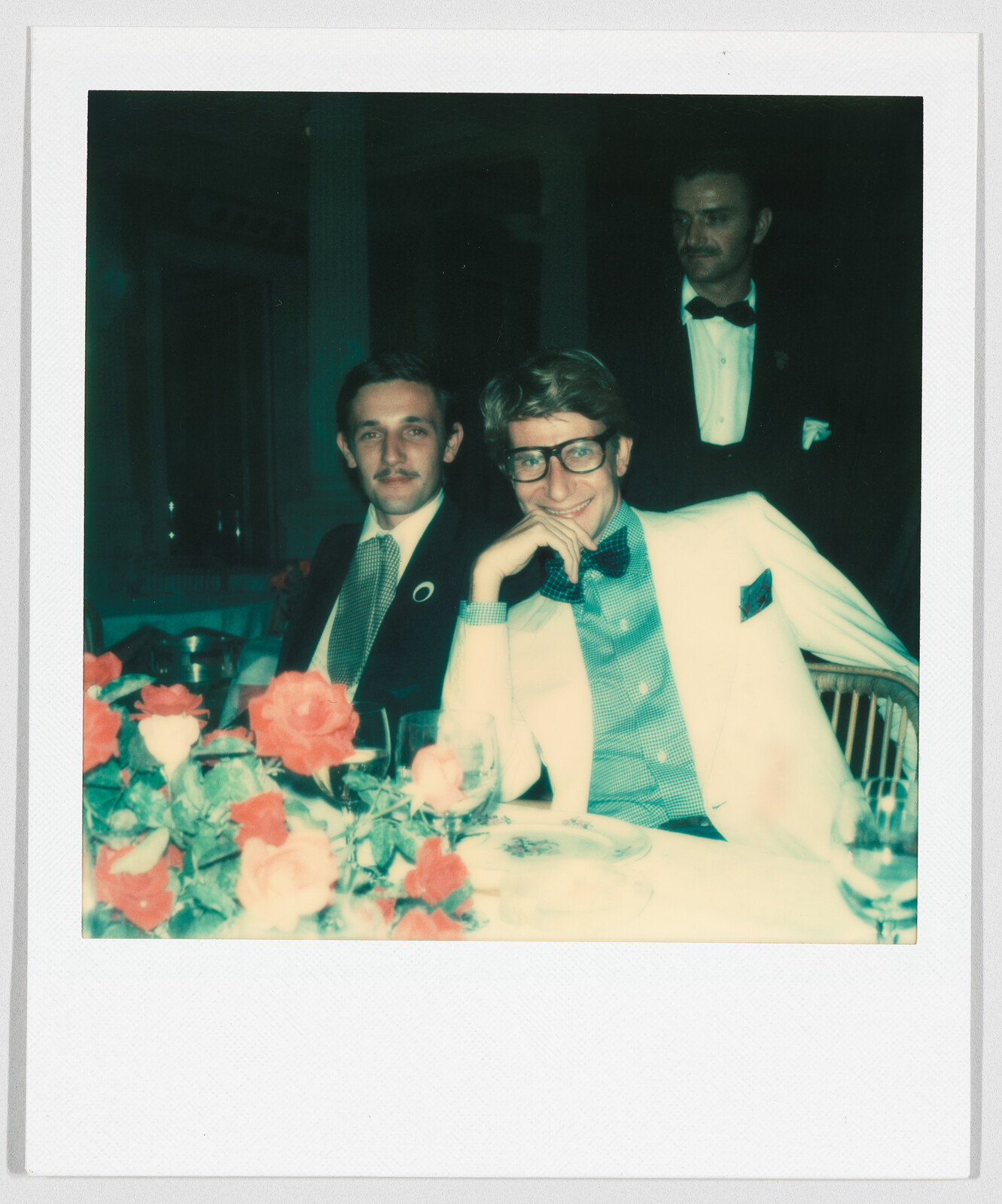 Two men in suits sit at a table with red flowers, smiling, while a waiter stands behind them.